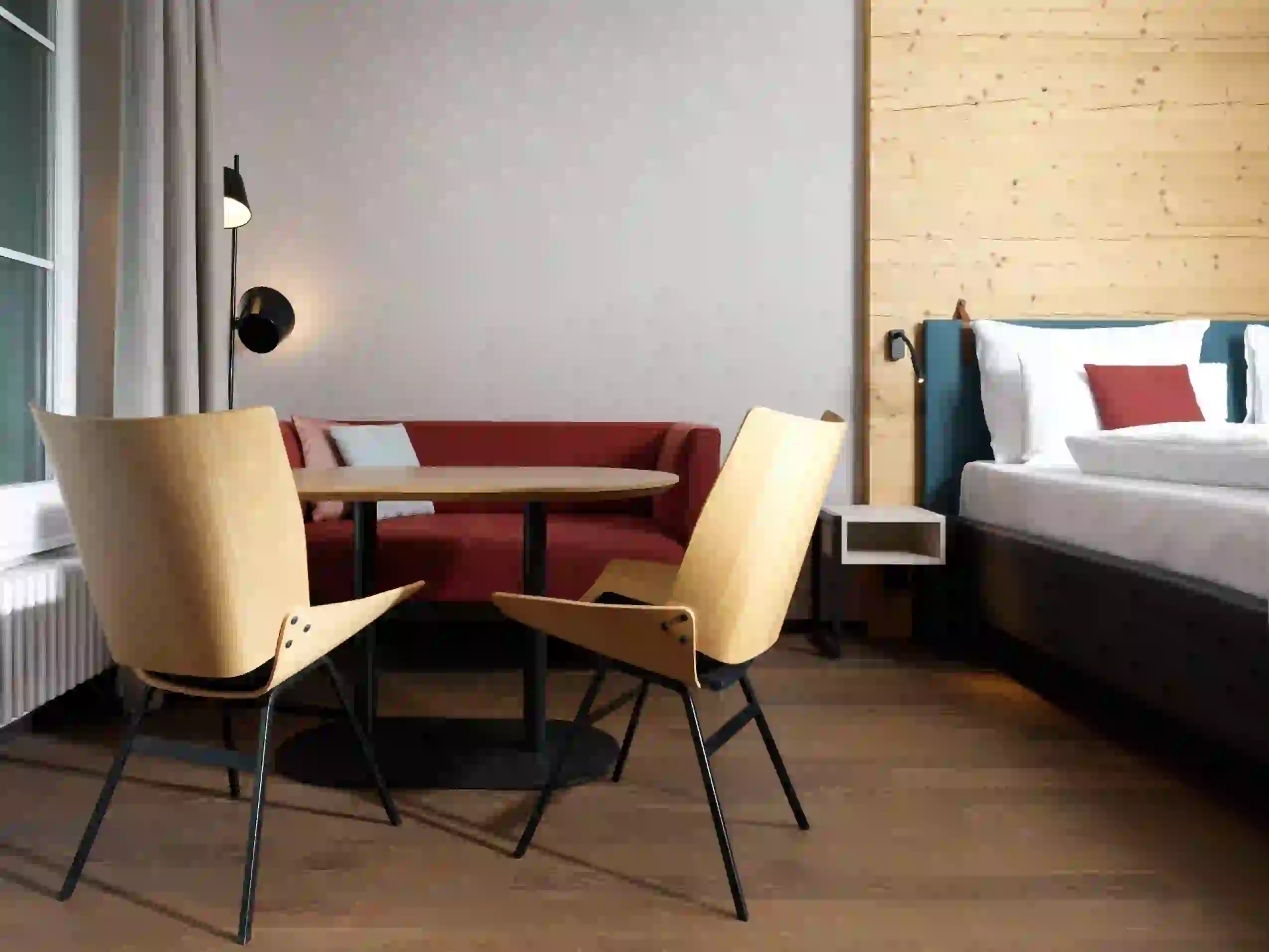 Ein modernes Hotelzimmer mit Holzboden, einem roten Sofa, einem runden Tisch mit zwei Holzstühlen, einem Bett mit weißer Bettwäsche und hellen holzgetäfelten und grauen Wänden.