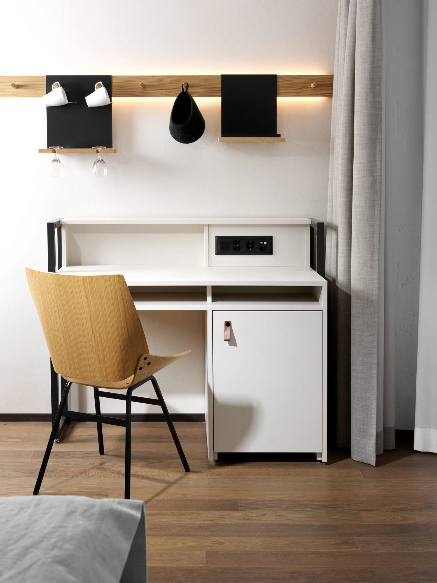 Ein moderner Arbeitsplatz im Hotel Jnterlaken mit Holzstuhl, weißem Schreibtisch mit Schrank, Steckdosen und Regal. Darüber befindet sich ein Holzwandregal mit zwei schwarzen Organizern und Gläsern, sanft beleuchtet von hinten.