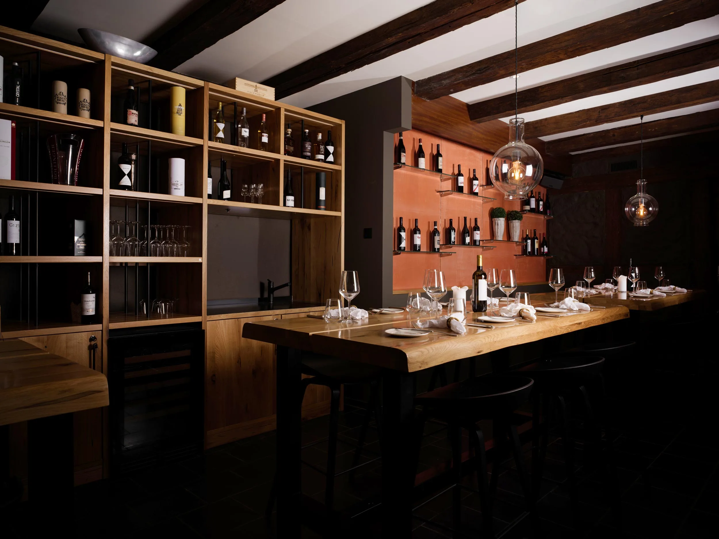 Eine gemütliche, schwach beleuchtete Weinbar mit Holzregalen voller Flaschen und Gläser, einem langen Holztisch mit Weingläsern, Tellern und Servietten und modernen Pendelleuchten, die von der Decke hängen.