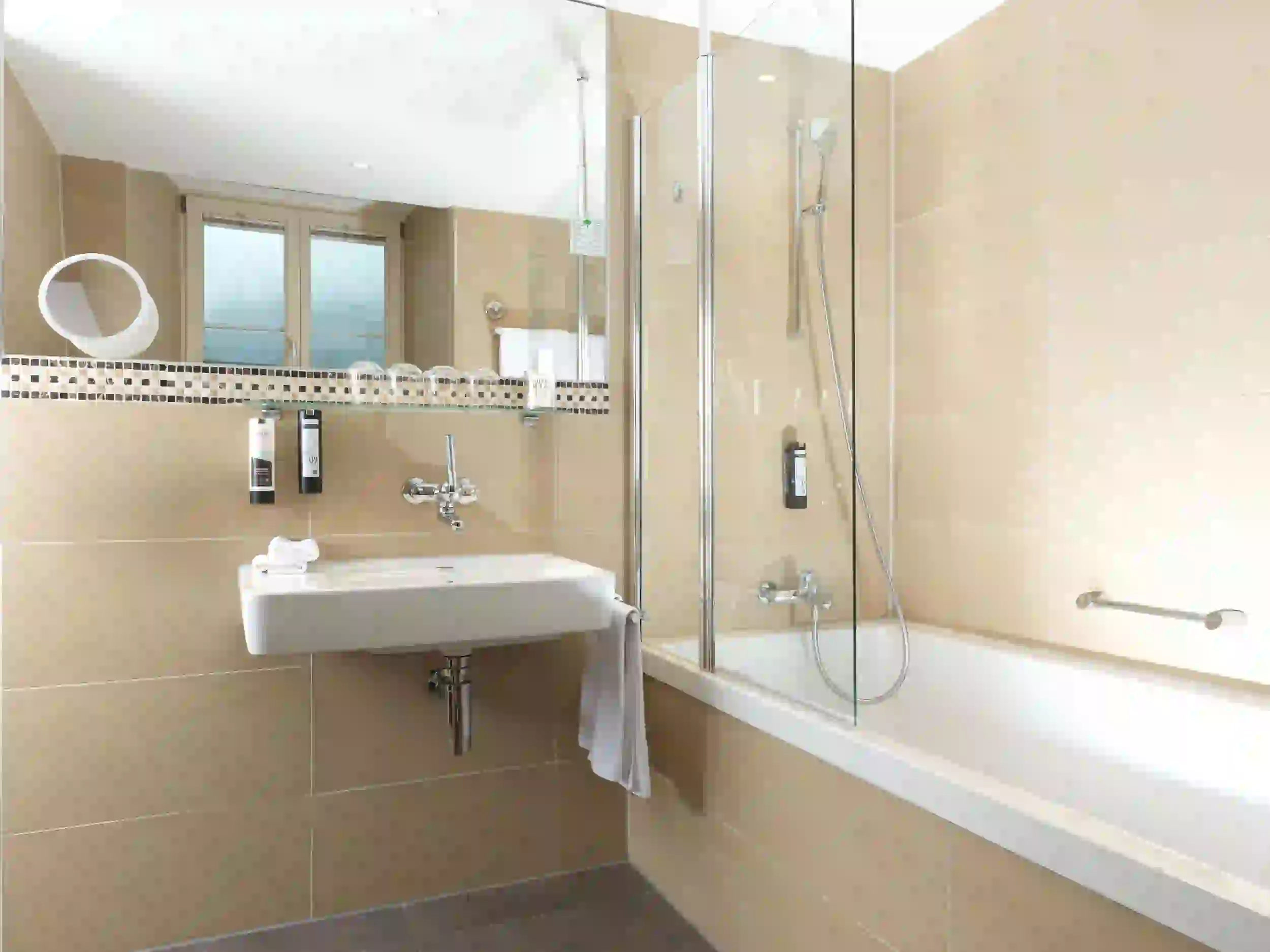 Modernes Badezimmer mit beigefarbenen Fliesen, einem schwebenden Waschbecken mit Handtuchhalter, einem Wandspiegel, einer Badewanne mit Glasduschwand und Pflegeprodukten. Große Fenster und helle Beleuchtung sorgen für ein klares, großzügiges Ambiente.