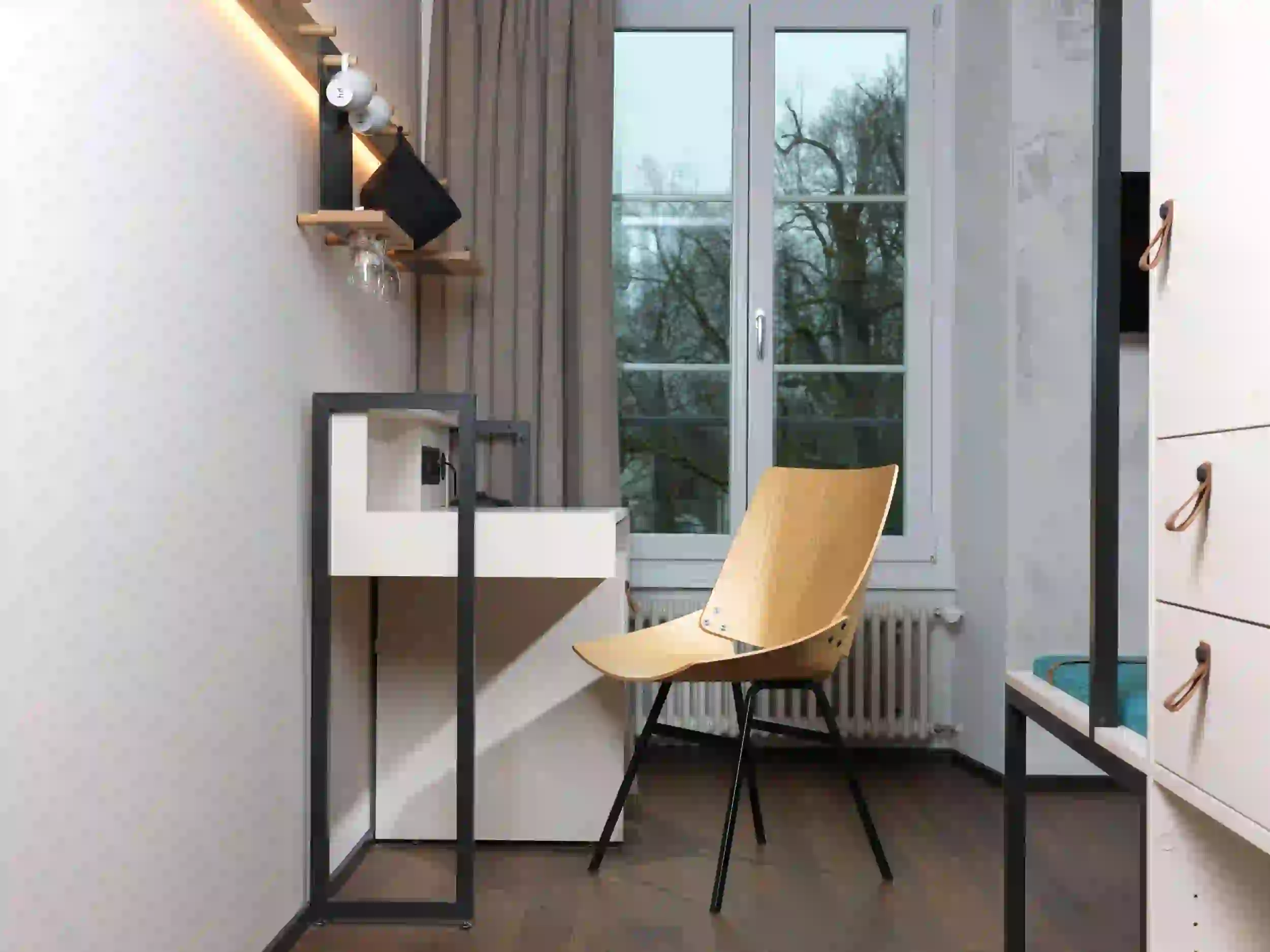 Minimalistischer Arbeitsbereich mit einem Holzstuhl, einem weißen Schreibtisch, an der Wand montierten Regalen mit Tassen und Gläsern und einem großen Fenster mit beigen Vorhängen mit Blick auf die kahlen Bäume draußen.
