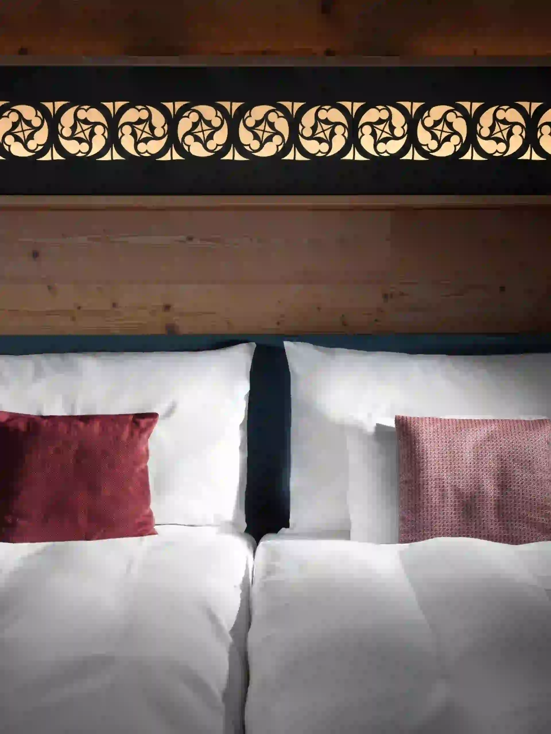 Ein ordentlich gemachtes Bett mit weißer Bettwäsche und zwei burgunderfarbenen Kissen vor einem hölzernen Kopfteil. Darüber befindet sich eine dekorative Platte mit einem komplizierten, hinterleuchteten Wirbelmuster.