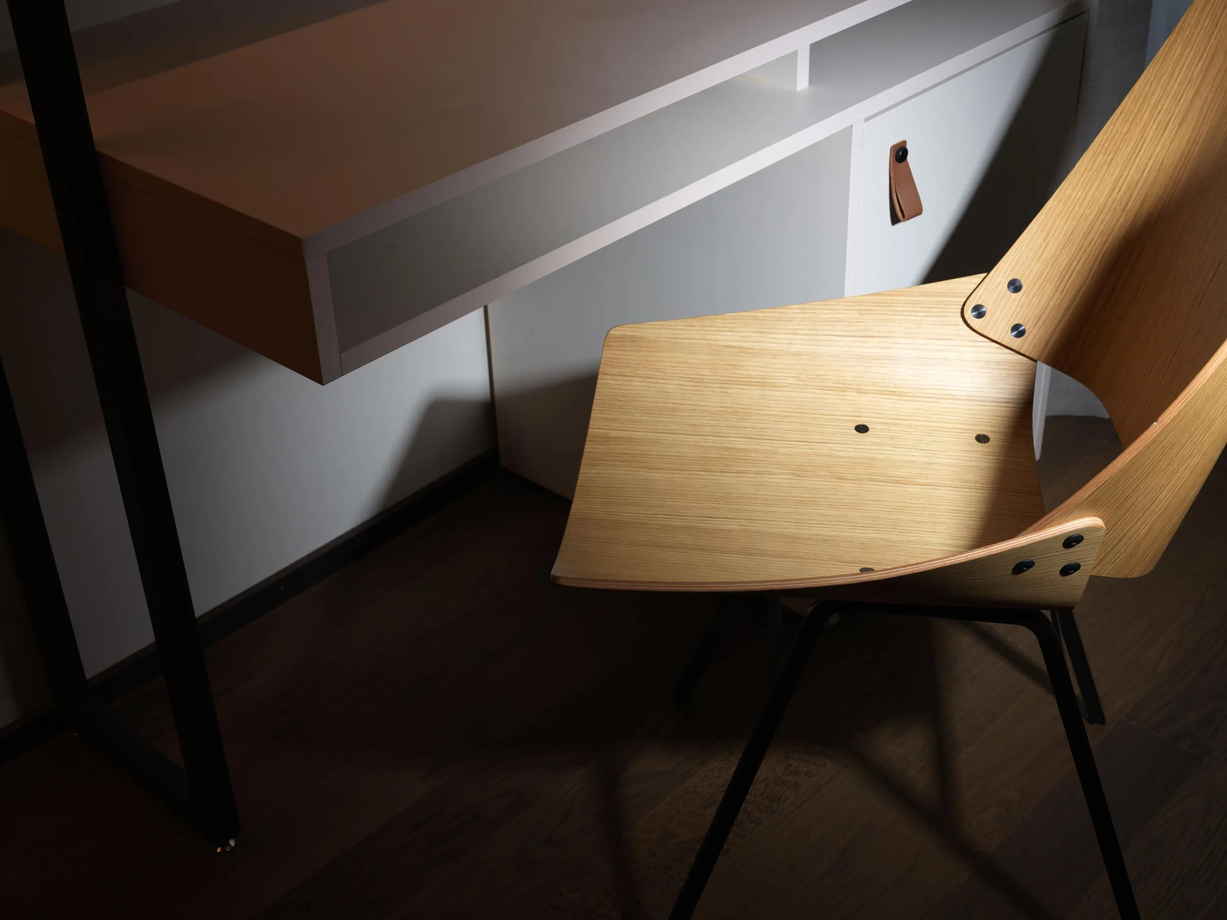 Ein moderner Holzstuhl mit schwarzen Metallbeinen steht neben einem minimalistischen weißen Schreibtisch mit Schublade auf einem dunklen Holzboden unter sanfter Beleuchtung.