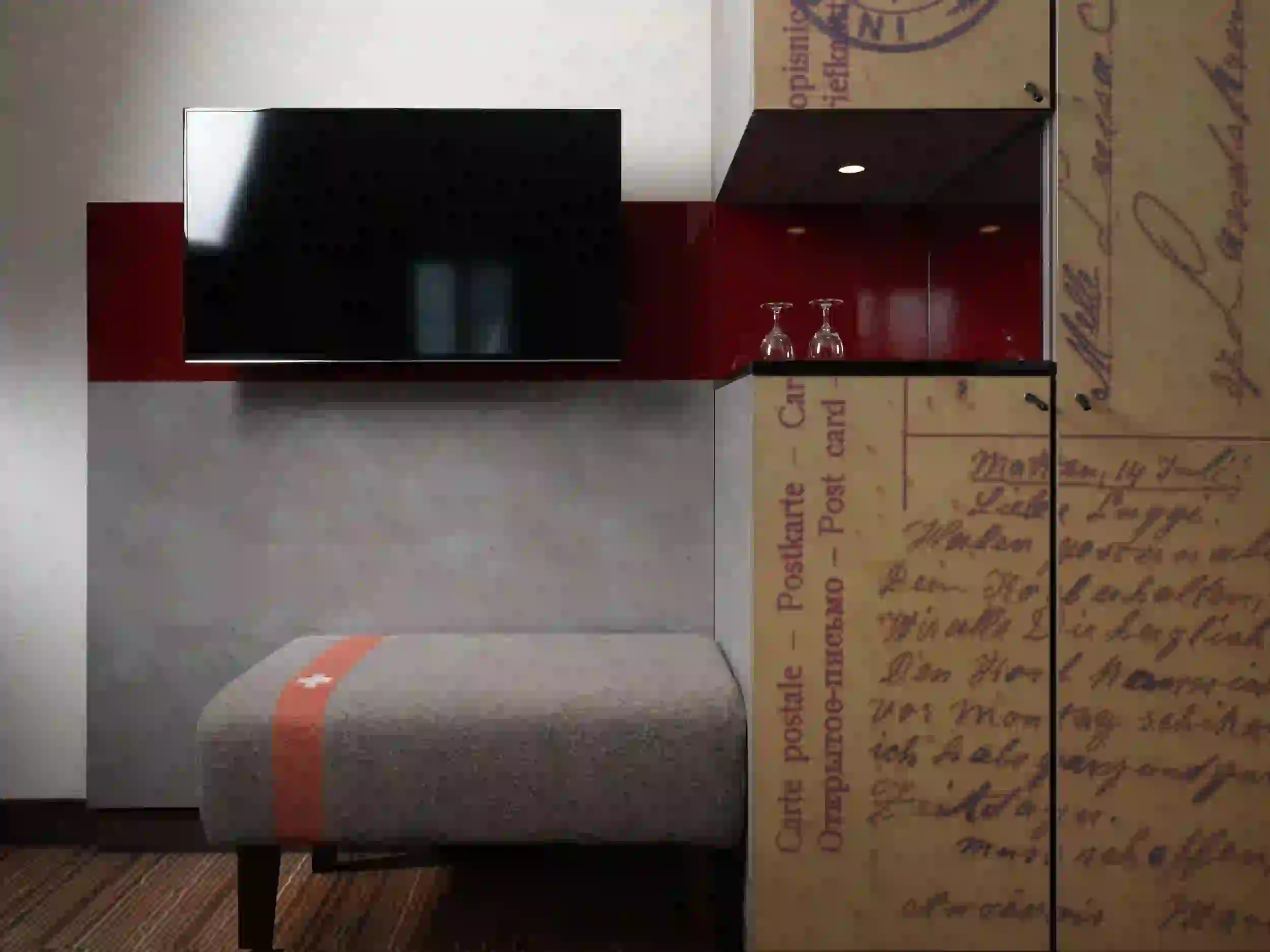 Eine moderne Hotelzimmerecke mit einem an der Wand montierten Fernseher über einer grauen Bank, roten und grauen Wandpaneelen und einem Schrank mit Vintage-Postkartendruck und zwei umgedrehten Gläsern auf einem dunklen Regal.
