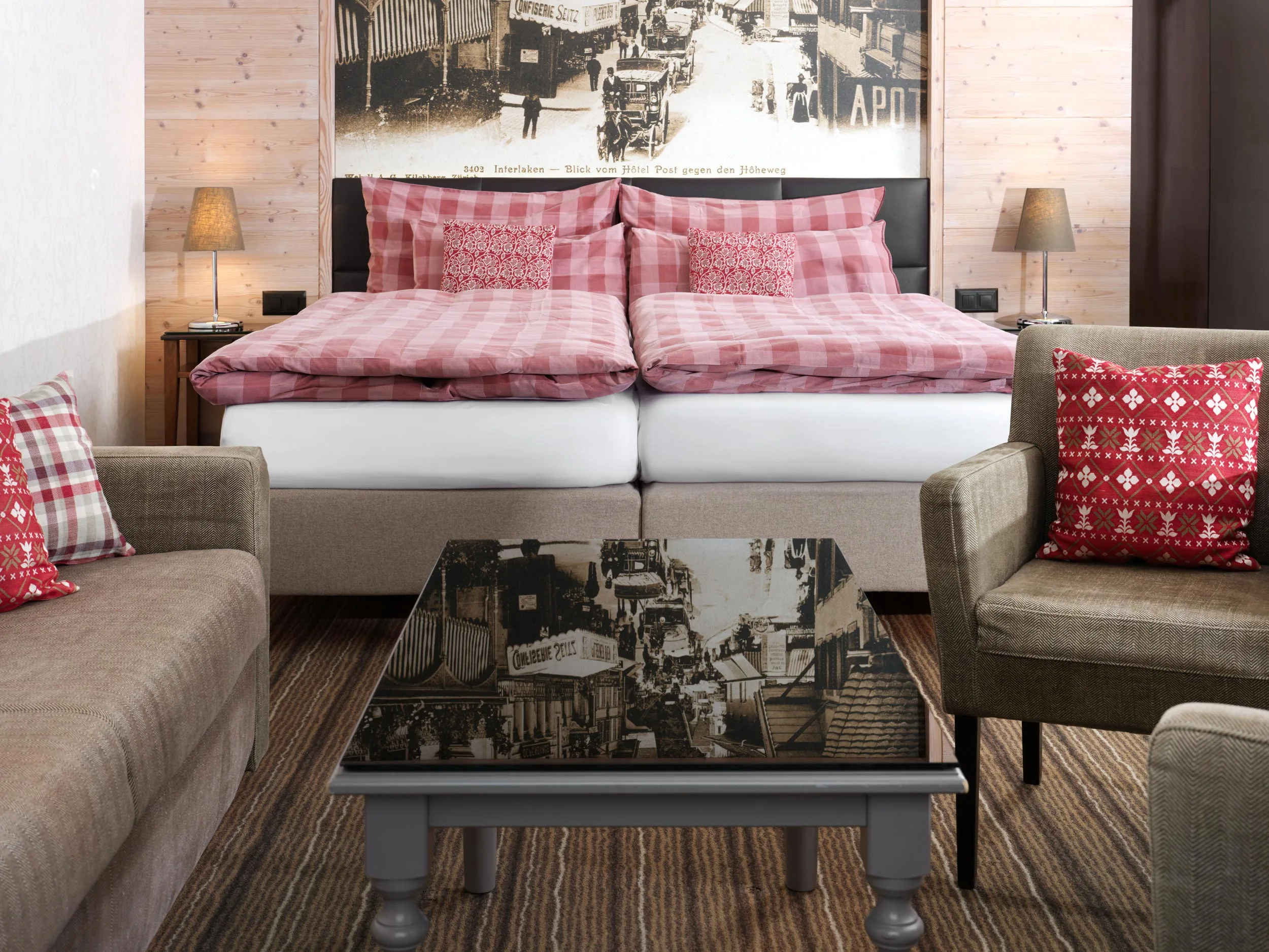 Ein gemütliches Hotelzimmer mit zwei Betten mit rot karierter Bettwäsche, passenden Kissen, einem braunen Sofa, einem Sessel mit einem rot gemusterten Kissen und einem Couchtisch aus Glas, an dessen Wand sich ein altes Stadtfoto spiegelt.