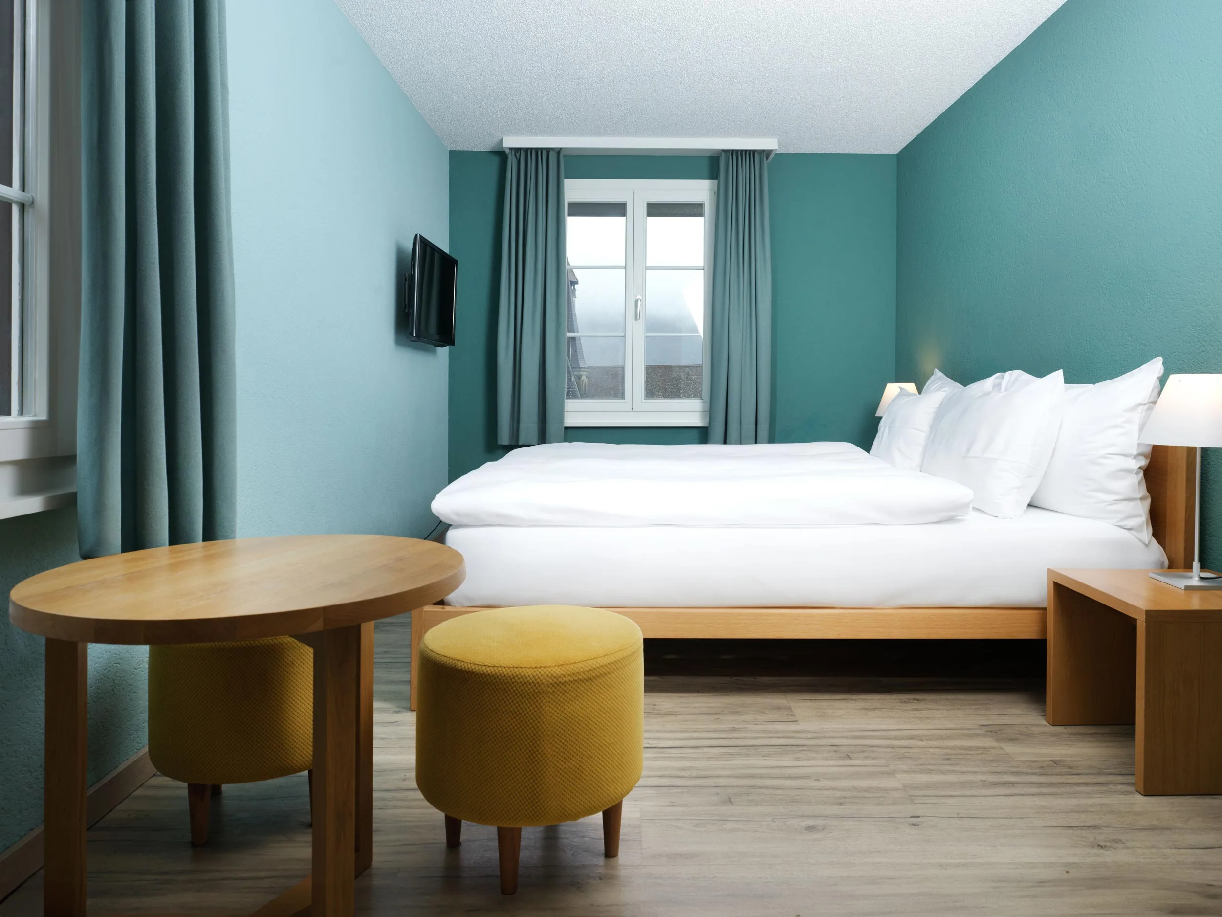 Ein modernes Hotelzimmer mit blaugrünen Wänden, einem großen Bett mit weißer Bettwäsche, einem Holztisch, zwei gelben Ottomanen, einer Nachttischlampe, einem Fernseher an der Wand und einem Fenster mit blaugrünen Vorhängen, die natürliches Licht hereinlassen.