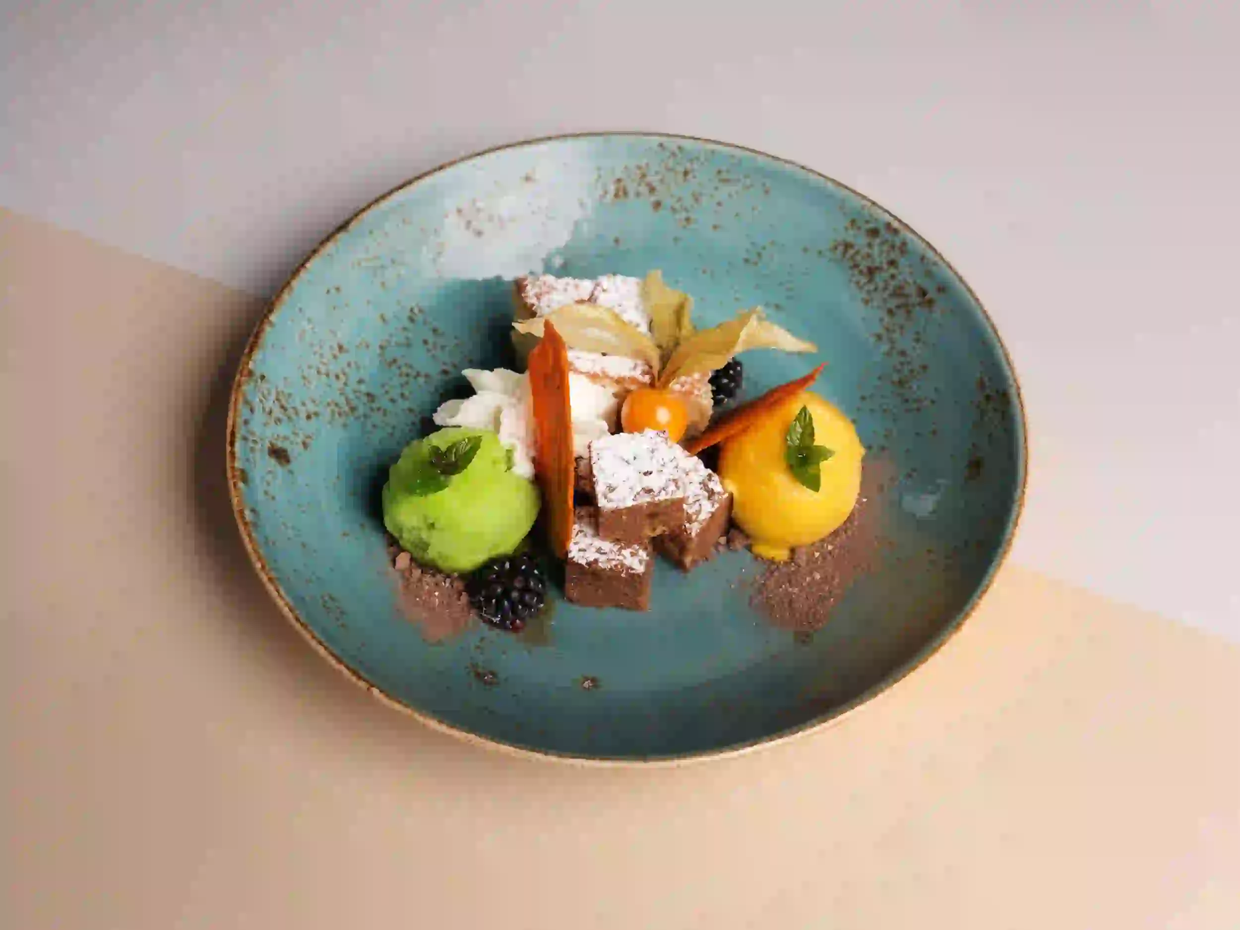 Ein blaugrüner Keramikteller mit kleinen Quadraten aus Puderzuckerkuchen, grünen und gelben Sorbetkugeln, Schlagsahne, Brombeeren, Schokoladenstreuseln und Garnituren, kunstvoll auf einem hellbeigen Hintergrund angeordnet.