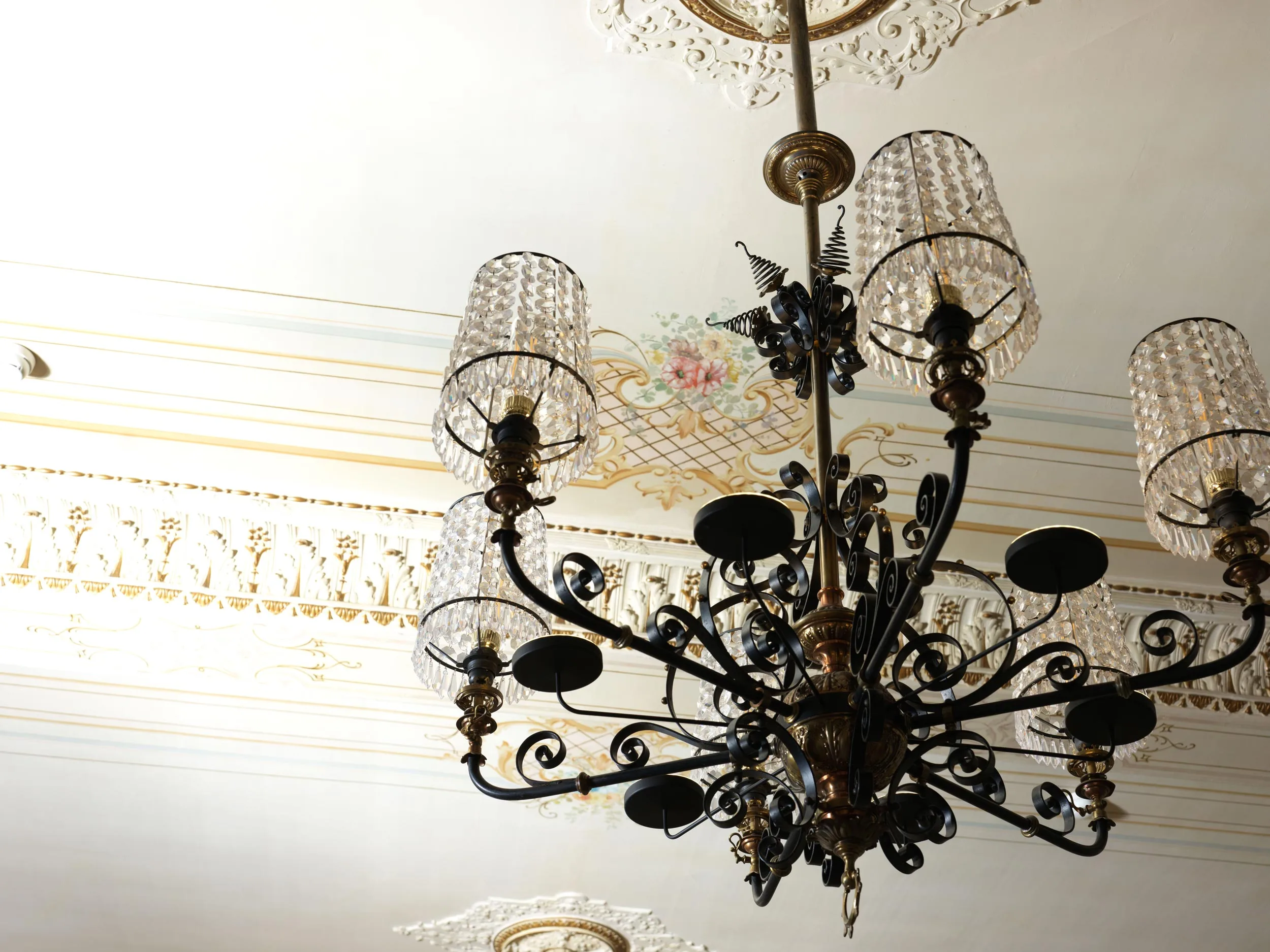 Ein verzierter schwarzer Kronleuchter mit sechs Lampenschirmen aus Glas hängt von einer kunstvoll verzierten weißen Decke mit Blumenmustern und komplizierten Verzierungen.