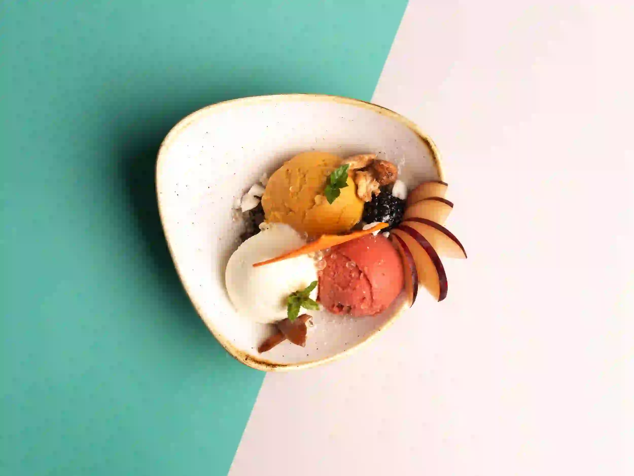 Eine Schüssel mit drei Kugeln buntem Eis, garniert mit Minze, Nüssen, Früchten und dünnen Pflaumenscheiben, auf einem zweifarbigen grün-rosa Hintergrund.