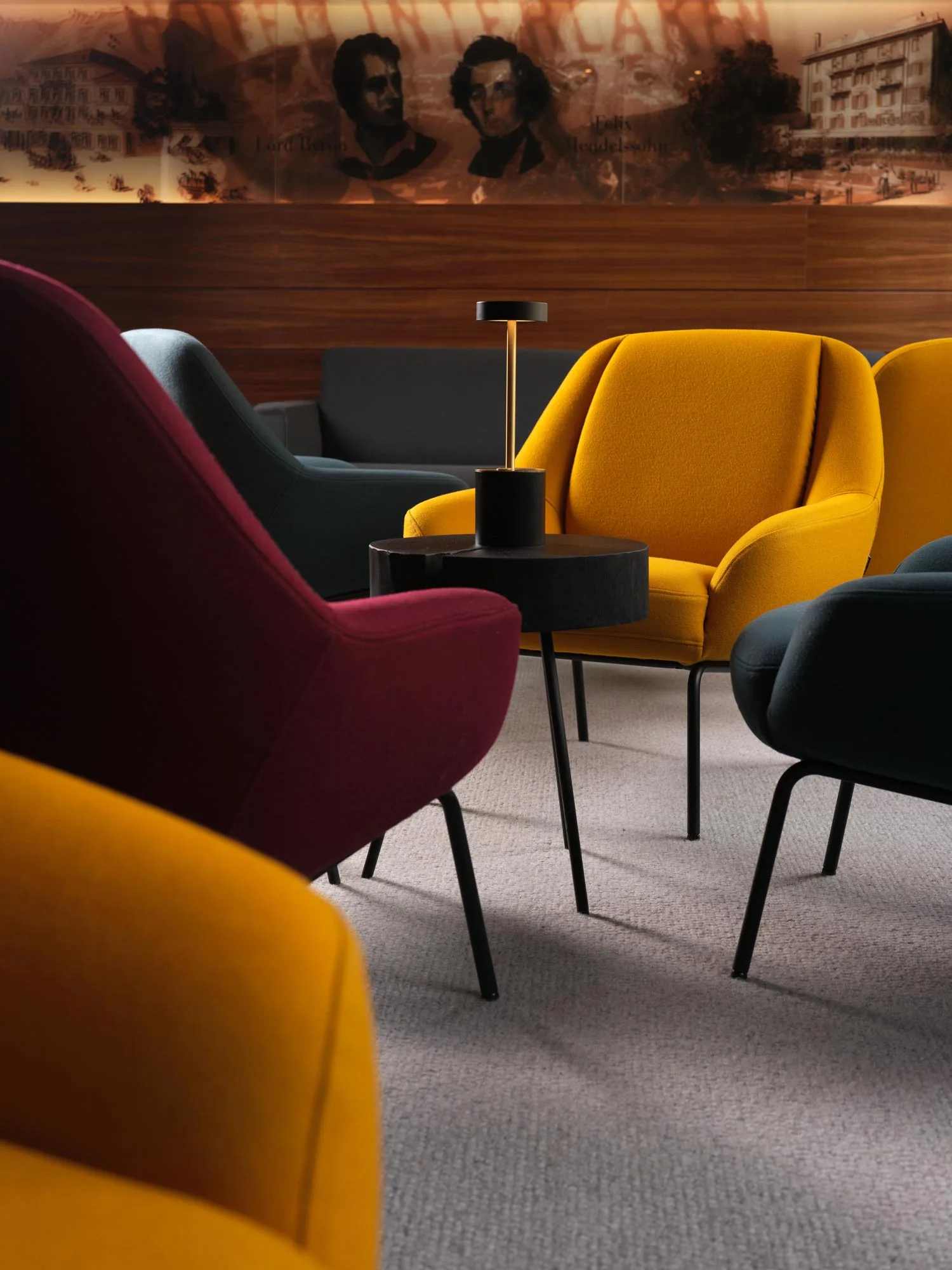 Moderner Loungebereich mit gelben, burgunderfarbenen und schwarzen Sesseln um kleine runde Tische, mit einer minimalistischen schwarzen Lampe und einer Wand mit historischen Porträts und Gebäuden in Sepia im Hintergrund.