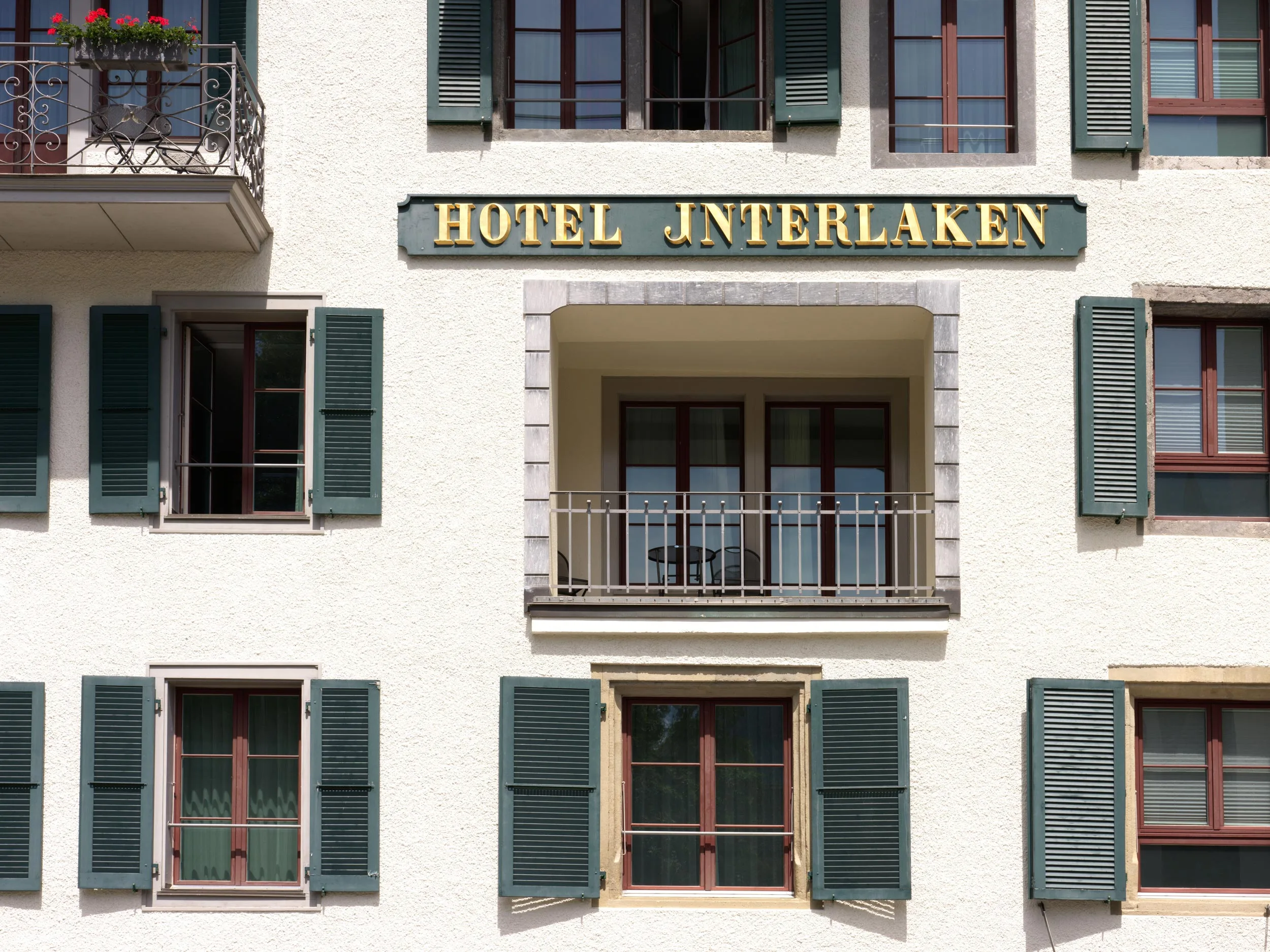 Die Fassade eines Gebäudes mit grünen Fensterläden, mehreren Fenstern und einem Balkon. Über dem Balkon steht in großen goldenen Buchstaben „HOTEL INTERLAKEN“.