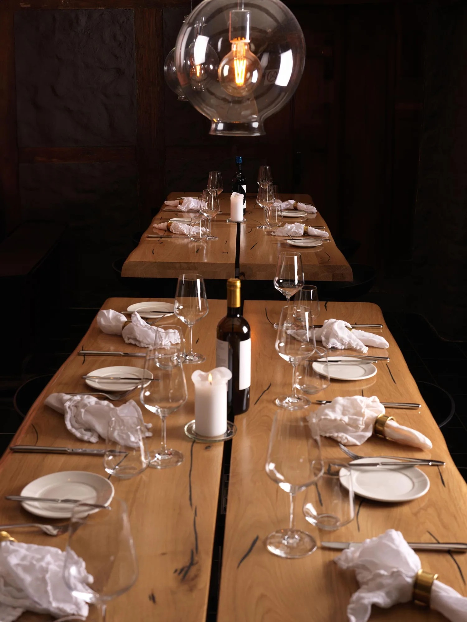 Ein für eine Mahlzeit gedeckter Esstisch aus Holz mit Weingläsern, weißen Servietten, Tellern, Kerzen und einer Weinflasche unter einer modernen Hängelampe in einem schwach beleuchteten Raum.