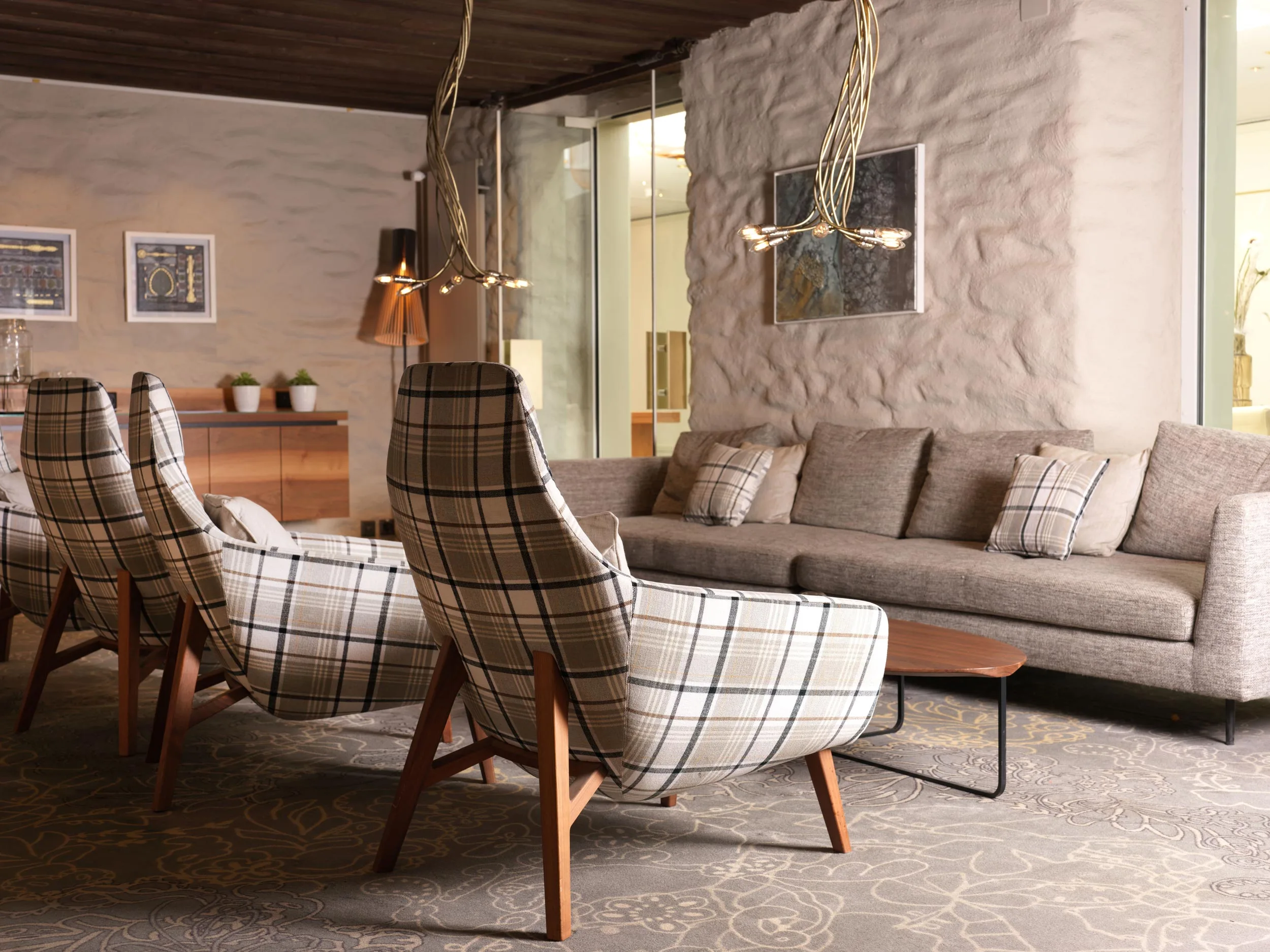 Moderner Loungebereich mit karierten Sesseln, einem grauen Sofa mit passenden Kissen, einem hölzernen Couchtisch und einzigartigen Pendelleuchten. Der Raum besticht durch strukturierte Wände, gerahmte Kunst und einen gemusterten Teppich.