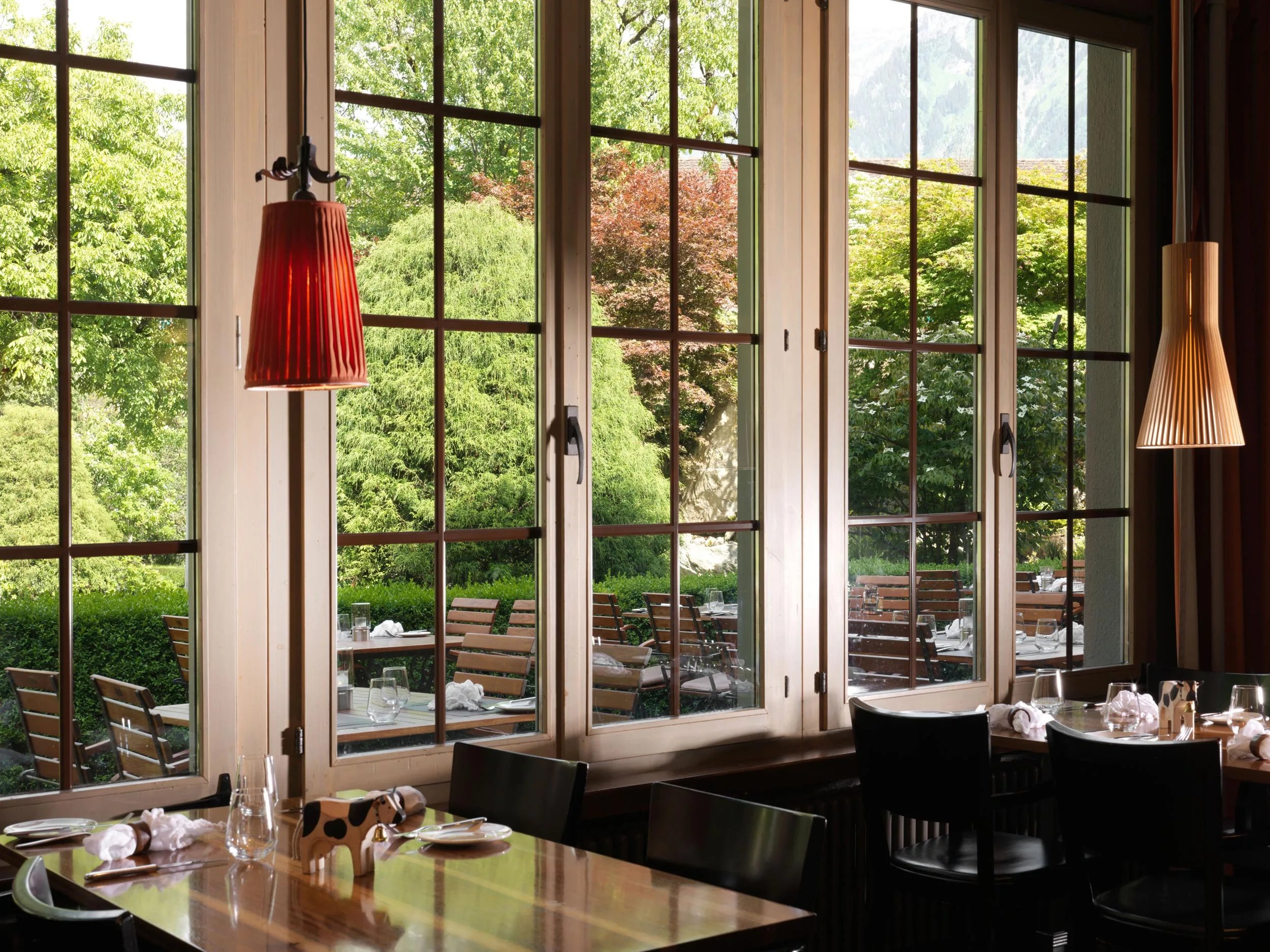 Ein Restaurant mit Holztischen und schwarzen Stühlen ist durch große Fenster erreichbar, die bei Tageslicht einen Blick auf Sitzgelegenheiten im Freien bieten, die von üppigen grünen Bäumen und Laub umgeben sind.