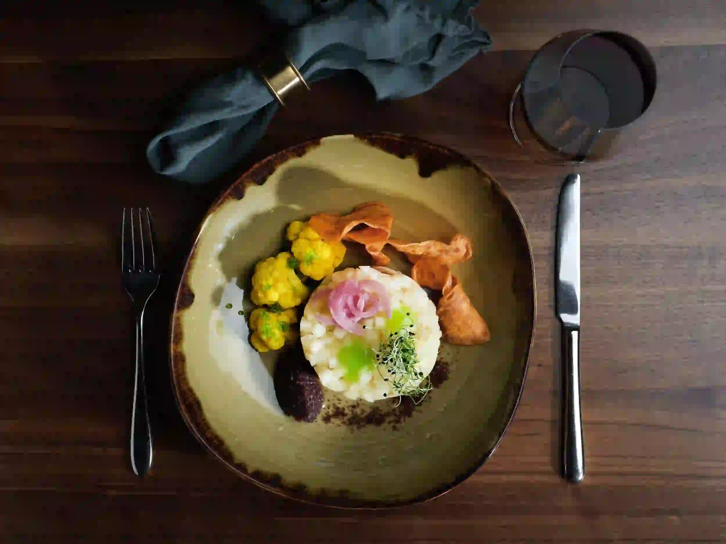 Ein Gourmet-Menü auf dem Teller mit Kartoffelpüree, eingelegten Zwiebeln, gelbem Blumenkohl, Süßkartoffelchips, einem dunklen Püree und Microgreens, serviert mit einem Glas Rotwein auf einem Holztisch mit Gabel, Messer und Serviette.