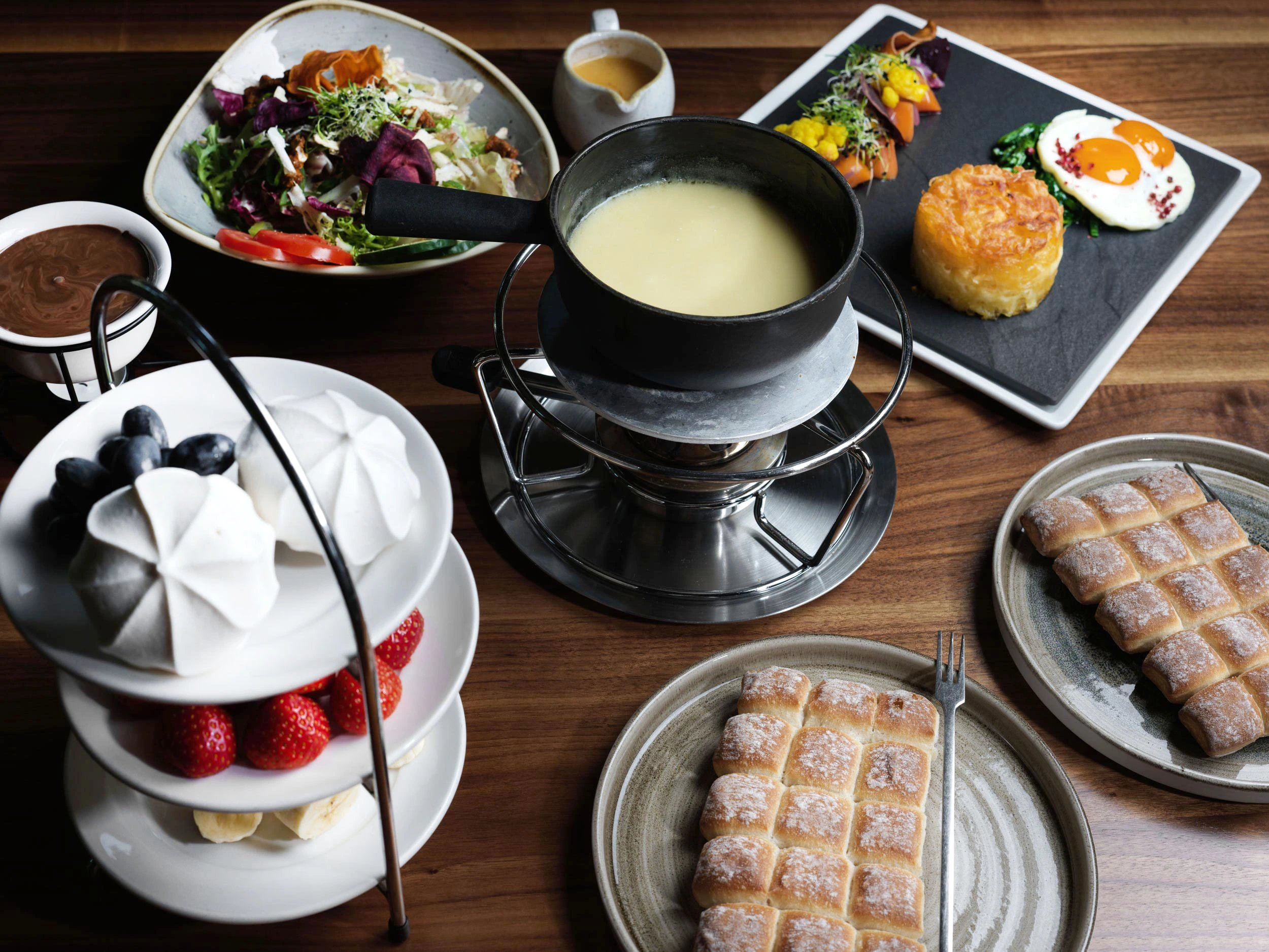 Ein gedeckter Tisch mit Käsefondue, einem frischen Salat, gegrilltem Gemüse mit Spiegelei, einem mehrstöckigen Tablett mit Baisers, Erdbeeren, Blaubeeren, Bananenscheiben und Tellern mit Waffelstangen und Schokoladendip.