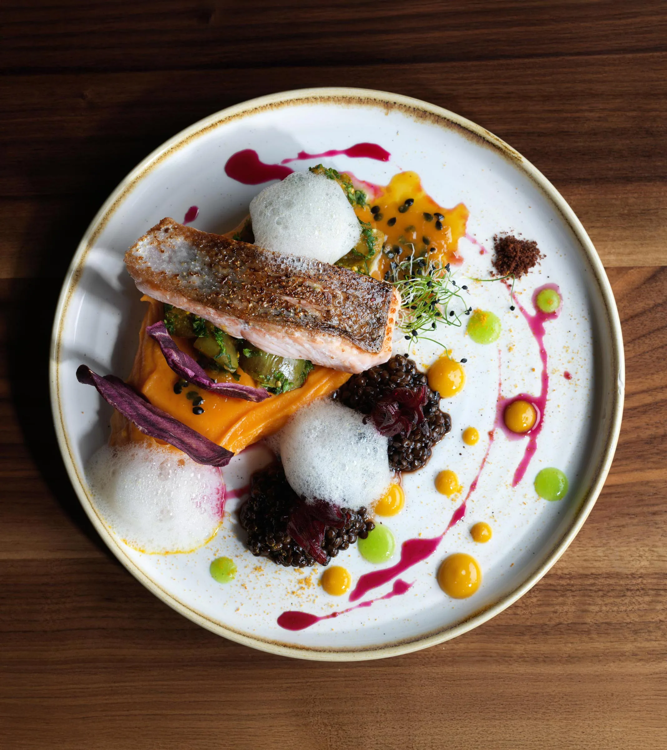 Ein Gourmetgericht auf dem Teller besteht aus einem gebratenen Lachsfilet auf Süßkartoffelpüree, Schaumgarnitur, schwarzem Quinoa, Mikrogrün und bunten Püree-Punkten, kunstvoll auf einem weißen Teller auf einem Holztisch angerichtet.