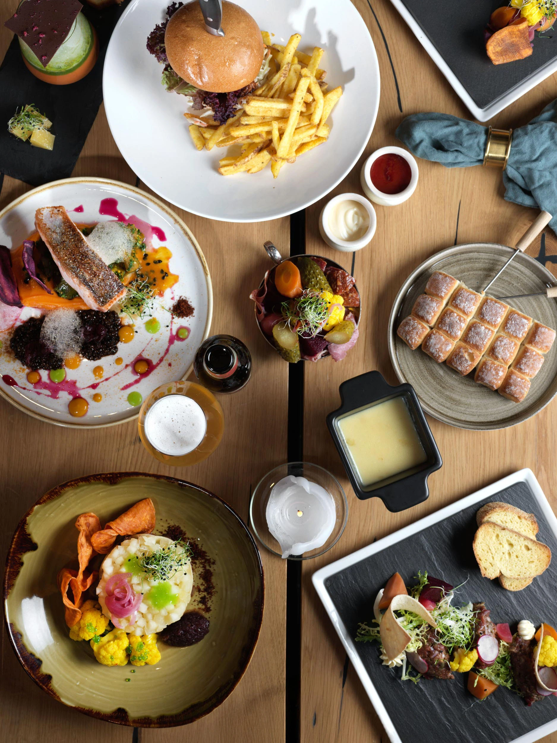 Draufsicht auf einen Holztisch mit verschiedenen Gerichten, darunter ein Burger mit Pommes frites, Lachs mit Soße, ein Käsefondue, geschnittenes Brot, Gemüse und Getränke. Die Teller sind fein säuberlich um den Tisch herum angeordnet.