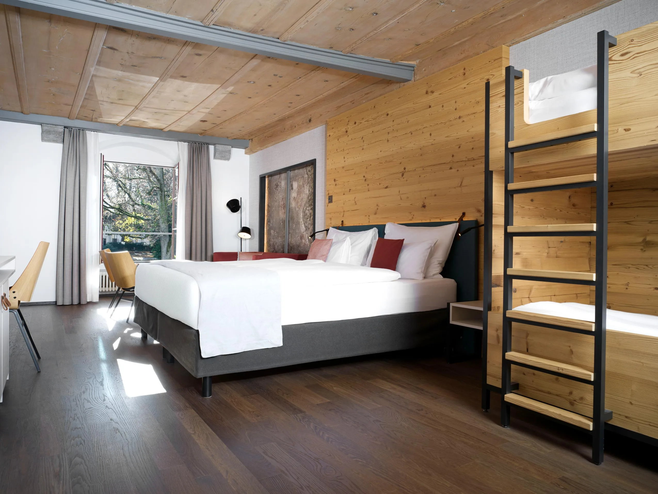 Ein modernes Hotelzimmer mit Holzwänden und -decke, einem Doppelbett mit weißer Bettwäsche, einem Etagenbett mit Leiter, einem Schreibtisch, Stühlen, einem großen Fenster mit Vorhängen und Hartholzböden.