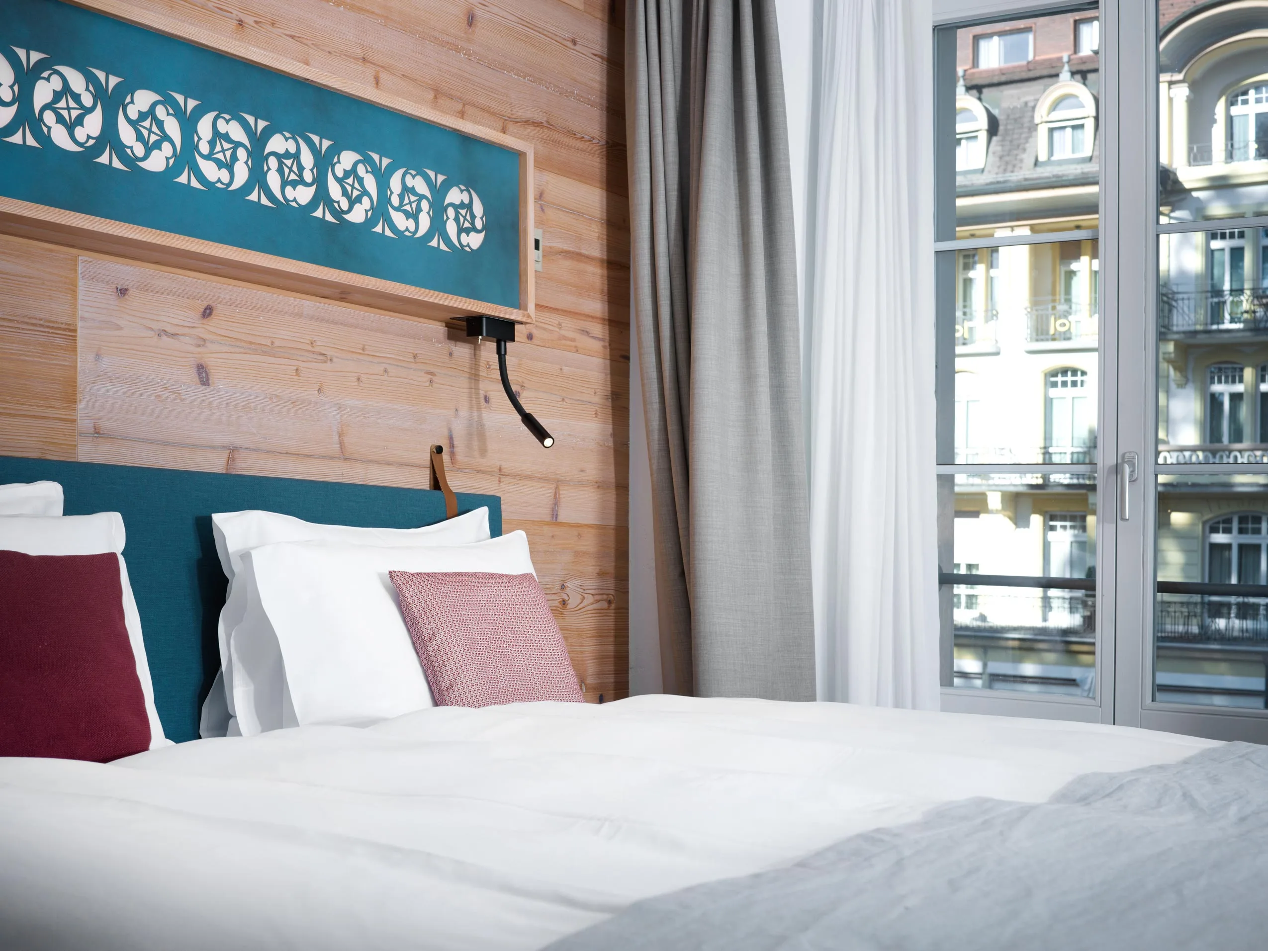 Ein ordentlich gemachtes Hotelzimmerbett mit weißer Bettwäsche und roten Kissen steht neben einem großen Fenster mit grauen Vorhängen und bietet Blick auf die Gebäude im klassischen europäischen Stil. Das Zimmer besticht durch helle Holzwände und moderne Einrichtung.