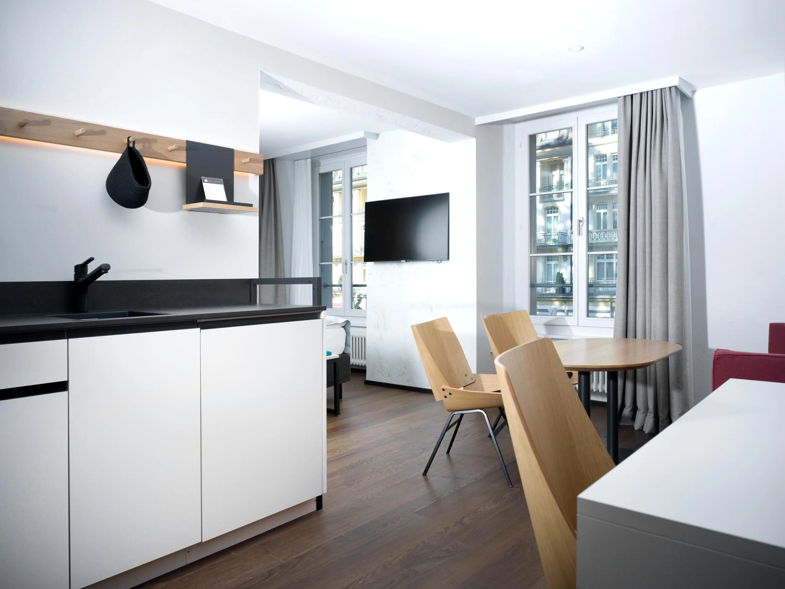 Modernes, helles Studio-Apartment mit kleiner Küche, rundem Esstisch und Holzstühlen, großen Fenstern mit grauen Vorhängen, an der Wand montiertem Fernseher und eleganten weißen Schränken – dieser Raum bietet den Komfort eines stilvollen Hotelzimmers mit viel Tageslicht.