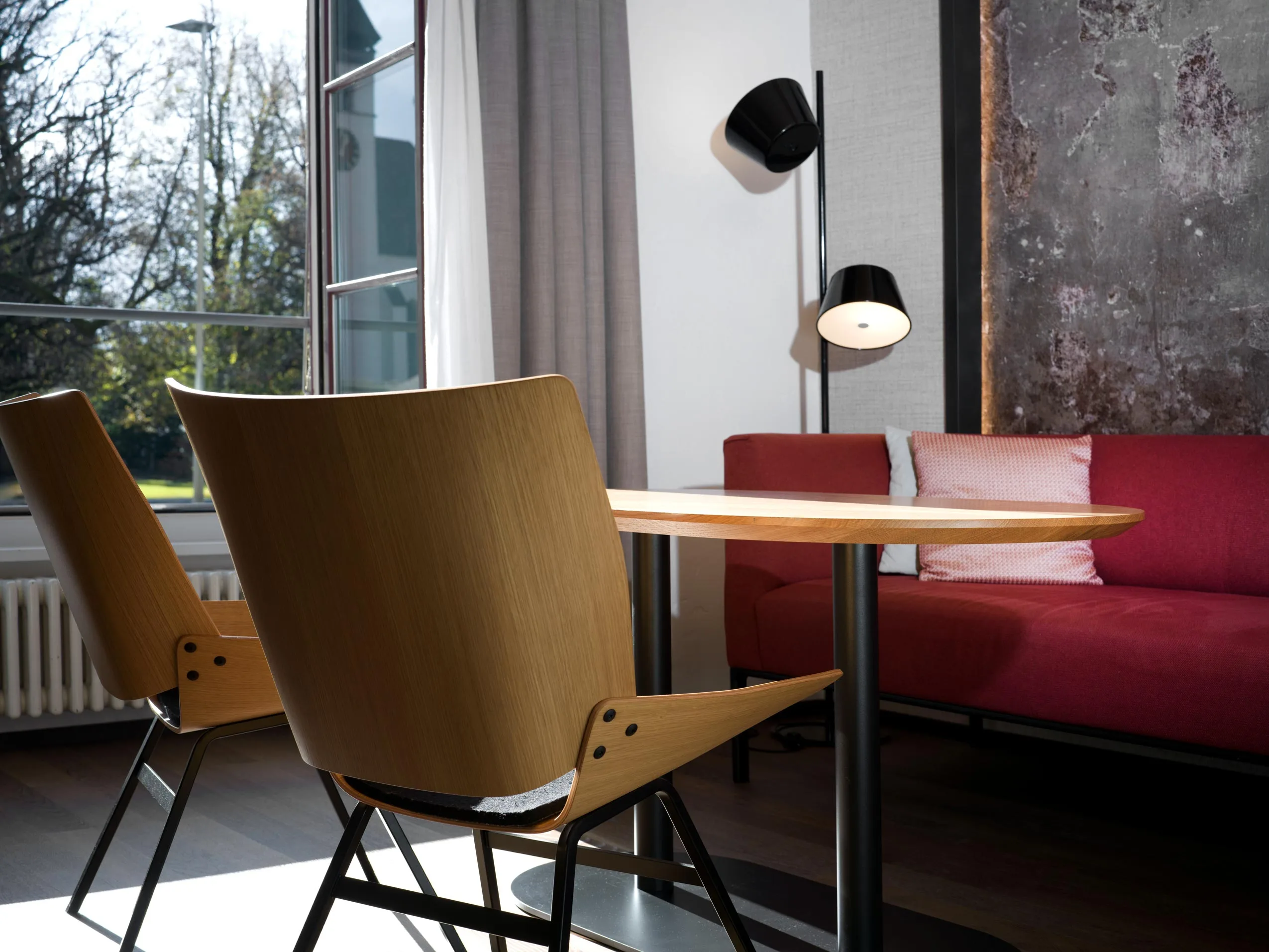 Modernes Wohnzimmer im Stil eines Hotelzimmers mit zwei Holzstühlen um einen hellen Holztisch, einem roten Sofa mit rosa Kissen, einer Stehlampe, einem großen Fenster mit Vorhängen und strukturierter Wanddekoration. Sonnenlicht fällt von draußen herein.