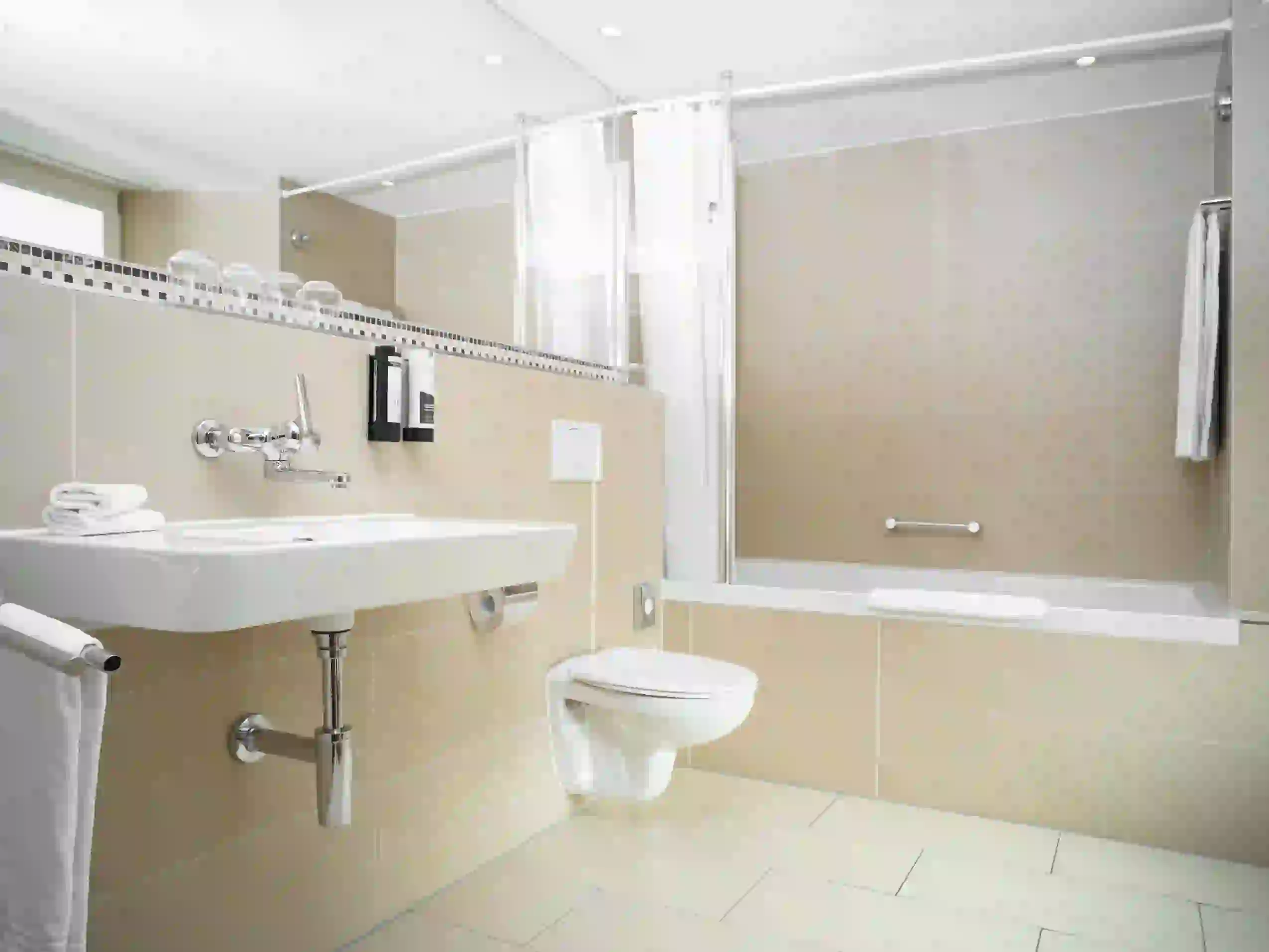 Modernes Hotelzimmerbad mit beigefarbenen Fliesen, Wandwaschbecken, WC und Badewanne mit weißem Duschvorhang. Handtücher liegen auf dem Waschbecken und hängen neben der Wanne. Glasablagen über dem Waschbecken sorgen für zusätzlichen Komfort.