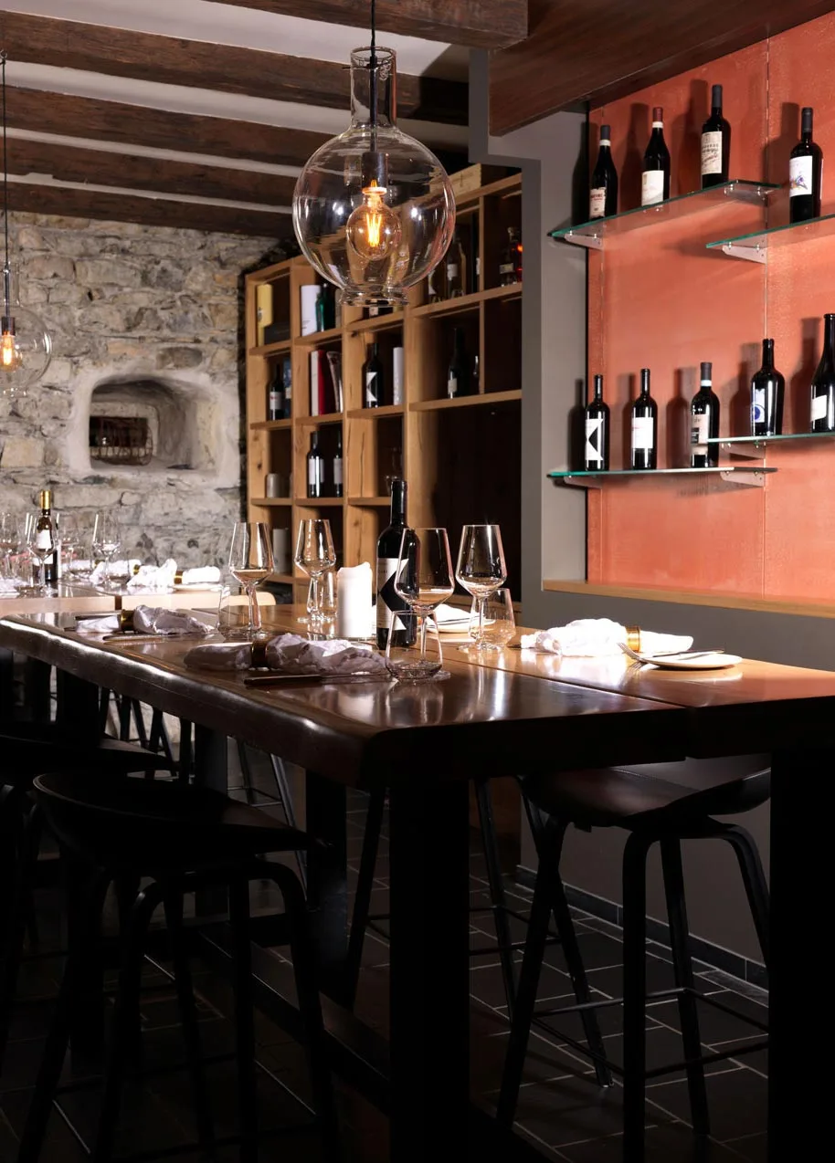 Ein stilvolles Restaurant-Interieur mit einem langen Holztisch zum Essen, modernen schwarzen Stühlen, hängenden Glasleuchten, Regalen mit Weinflaschen an der Wand und freiliegendem Mauerwerk im Hintergrund.