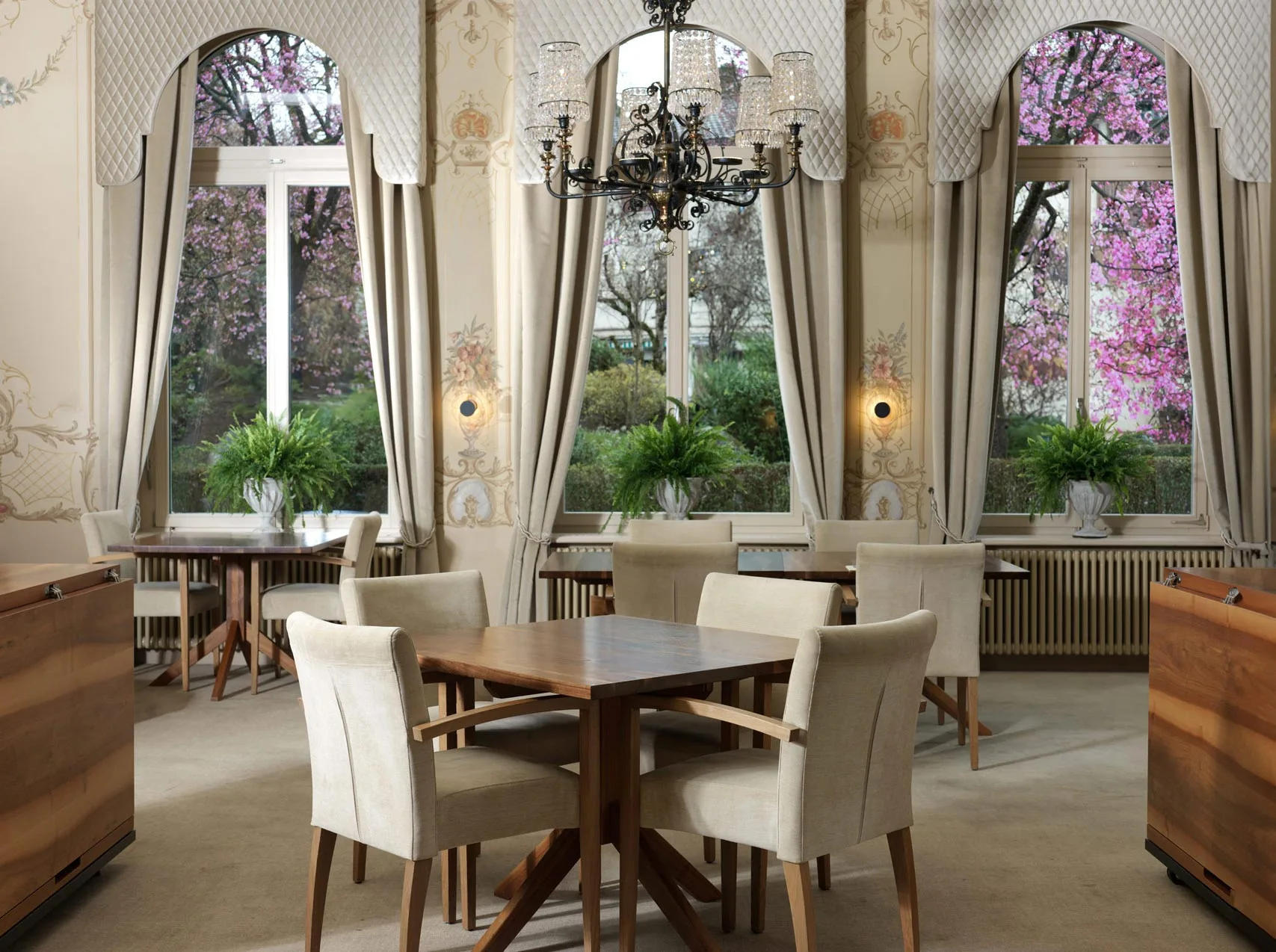 Elegantes Esszimmer mit beigen Stühlen und Holztischen, großen Fenstern mit cremefarbenen Vorhängen, dekorativem Kronleuchter, Topfpflanzen und Blick auf blühende Bäume im Freien. Sanfte Beleuchtung und kunstvolle Wandgestaltung sorgen für eine edle Atmosphäre.