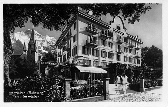 Schwarzweißfoto des historischen Hotels Interlaken in der Schweiz, mit einem Berg im Hintergrund, einer Kirche auf der linken Seite und zwei Personen, die in der Nähe des Hoteleingangs spazieren gehen, eingerahmt von Bäumen und einem schmiedeeisernen Zaun.