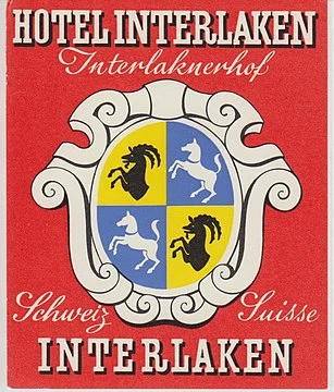Ein Hotelschild im Vintage-Stil zeigt ein dekoratives Wappen mit schwarz-weißen Pferden auf einem blau-gelben Schild und den Text „Hotel Interlaken“, „Schweiz Suisse“ und „Interlaken“ auf einem auffälligen roten Hintergrund.