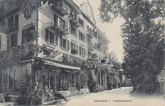 Ein altes Schwarzweißfoto des Interlaken Hotels, das ein mehrstöckiges Gebäude mit Balkonen, Fensterläden und Sitzgelegenheiten im Freien zeigt, umgeben von Bäumen und Grün.