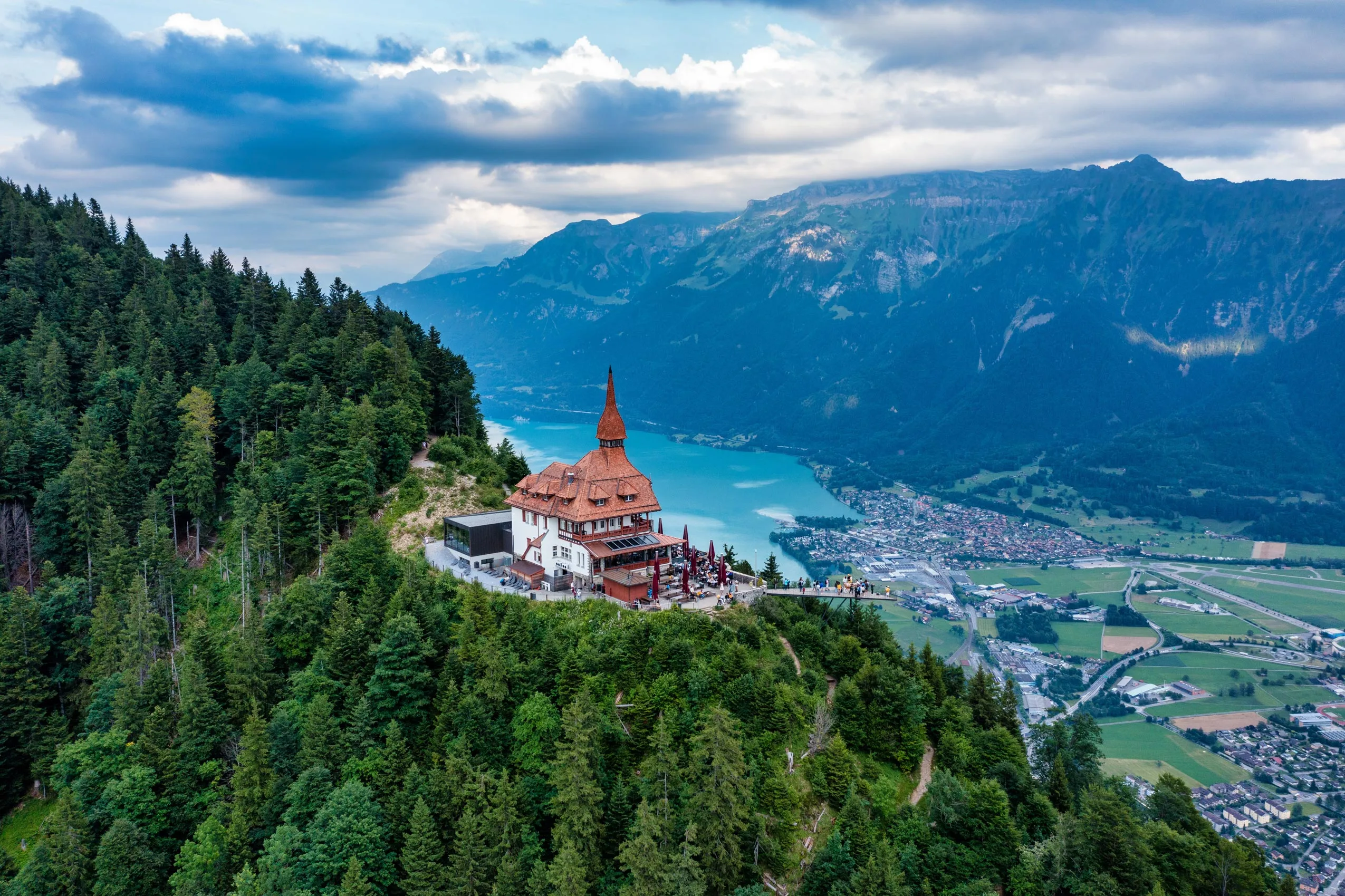 Ein großes Gebäude mit rotem Dach steht auf einem grünen, bewaldeten Hügel mit Blick auf einen türkisfarbenen See und eine Talstadt, mit dramatischen Bergen im Hintergrund unter einem bewölkten Himmel.
