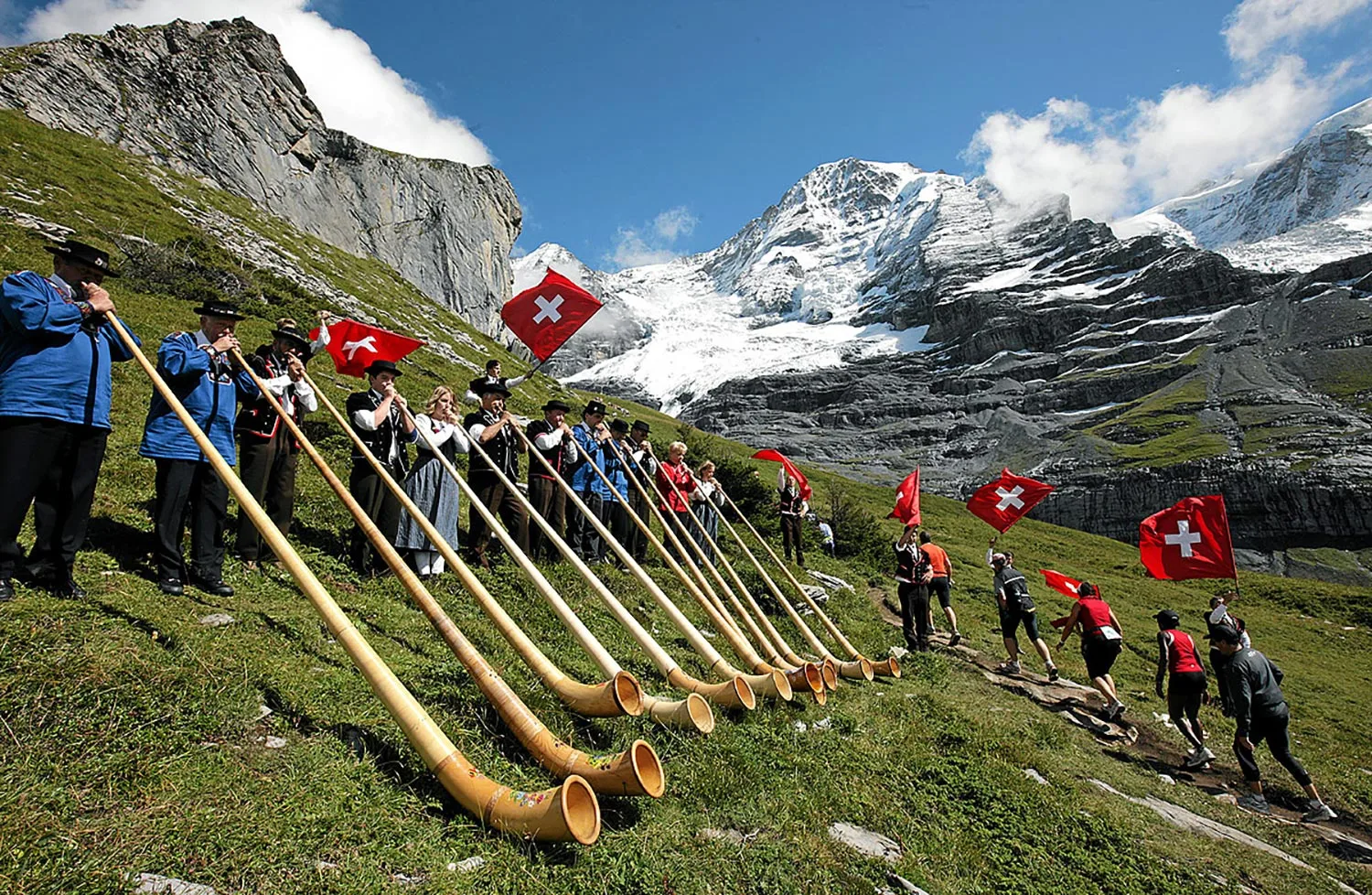 Eine Gruppe von Menschen in traditioneller Schweizer Kleidung spielt Alphörner auf einem grasbewachsenen Berghang, während andere Schweizer Fahnen halten. Im Hintergrund erheben sich schneebedeckte Berge unter einem teilweise bewölkten Himmel.