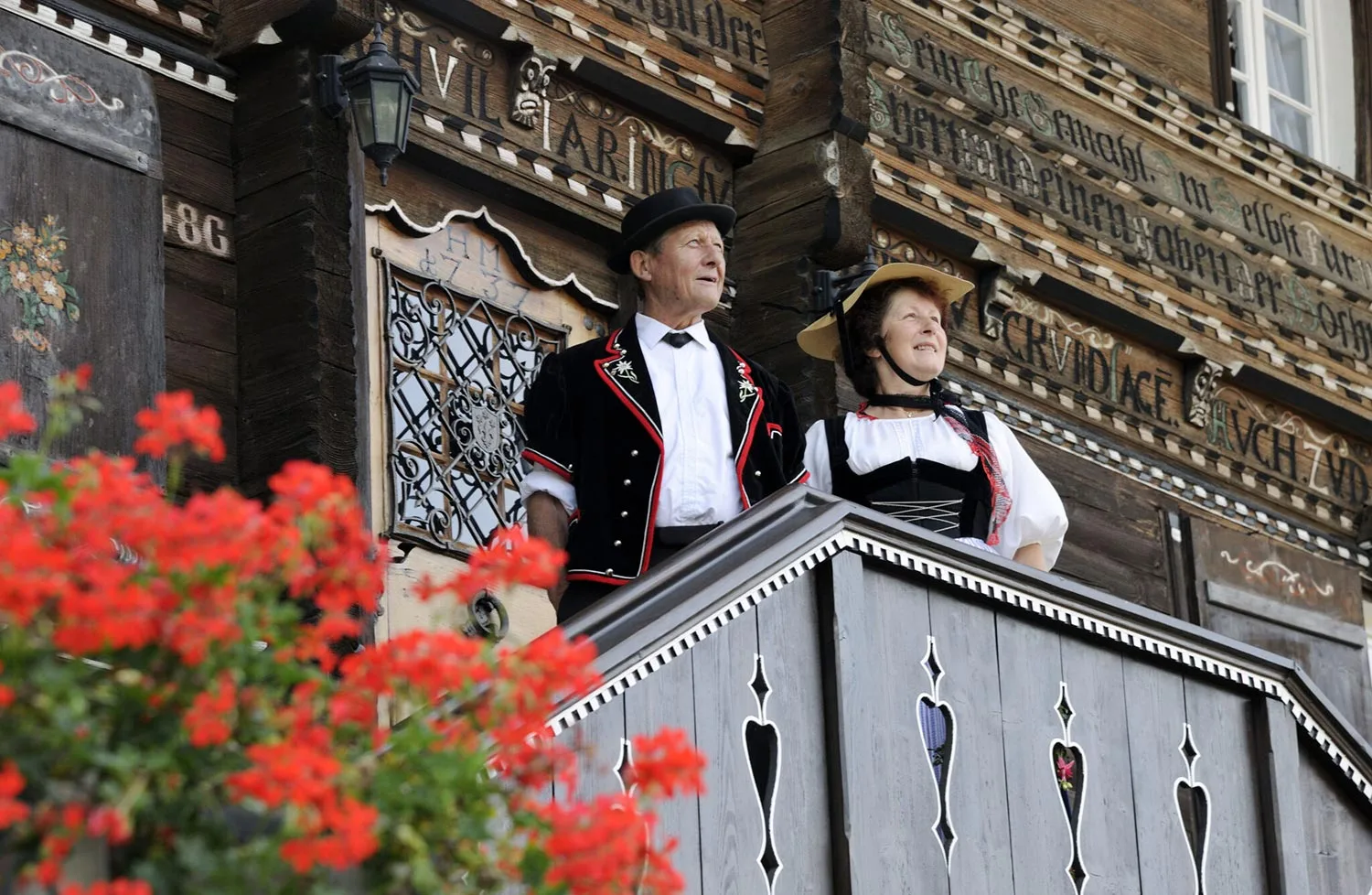 Ein Mann und eine Frau in traditioneller Schweizer Kleidung stehen auf einem mit Herzausschnitten verzierten Holzbalkon. Im Vordergrund sind rote Blumen zu sehen, und kunstvolle Schnitzereien schmücken das Gebäude dahinter.