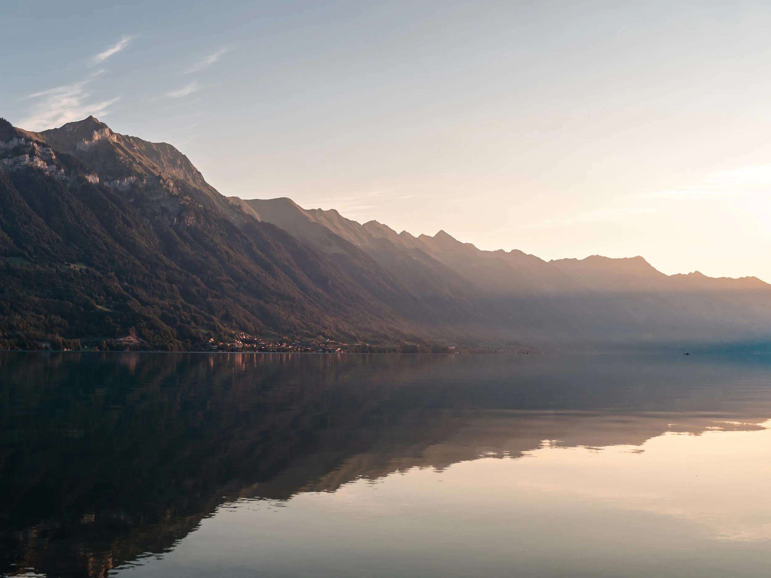 Ruhiger See bei Sonnenaufgang oder Sonnenuntergang mit im Wasser spiegelnden Bergen, getaucht in sanftes goldenes Licht. Etwas Dunst und sanfte Wolken am Himmel schaffen eine friedliche und heitere Landschaftsszene.