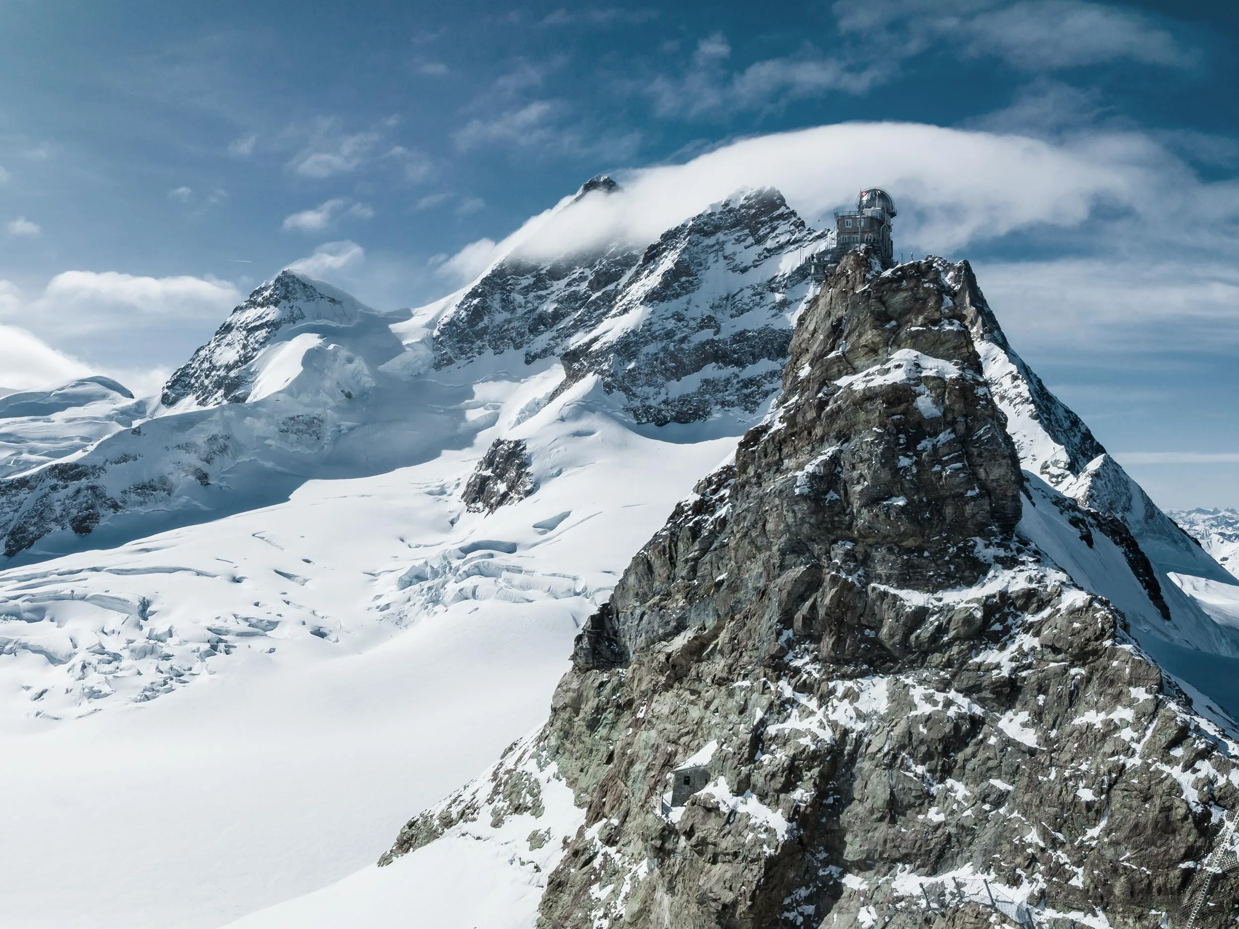 Schneebedeckte Berge unter einem blauen Himmel mit Wolken; im Vordergrund ist ein markanter Felsgipfel zu sehen, während sich im Hintergrund größere, abgerundete Schneegipfel erheben.