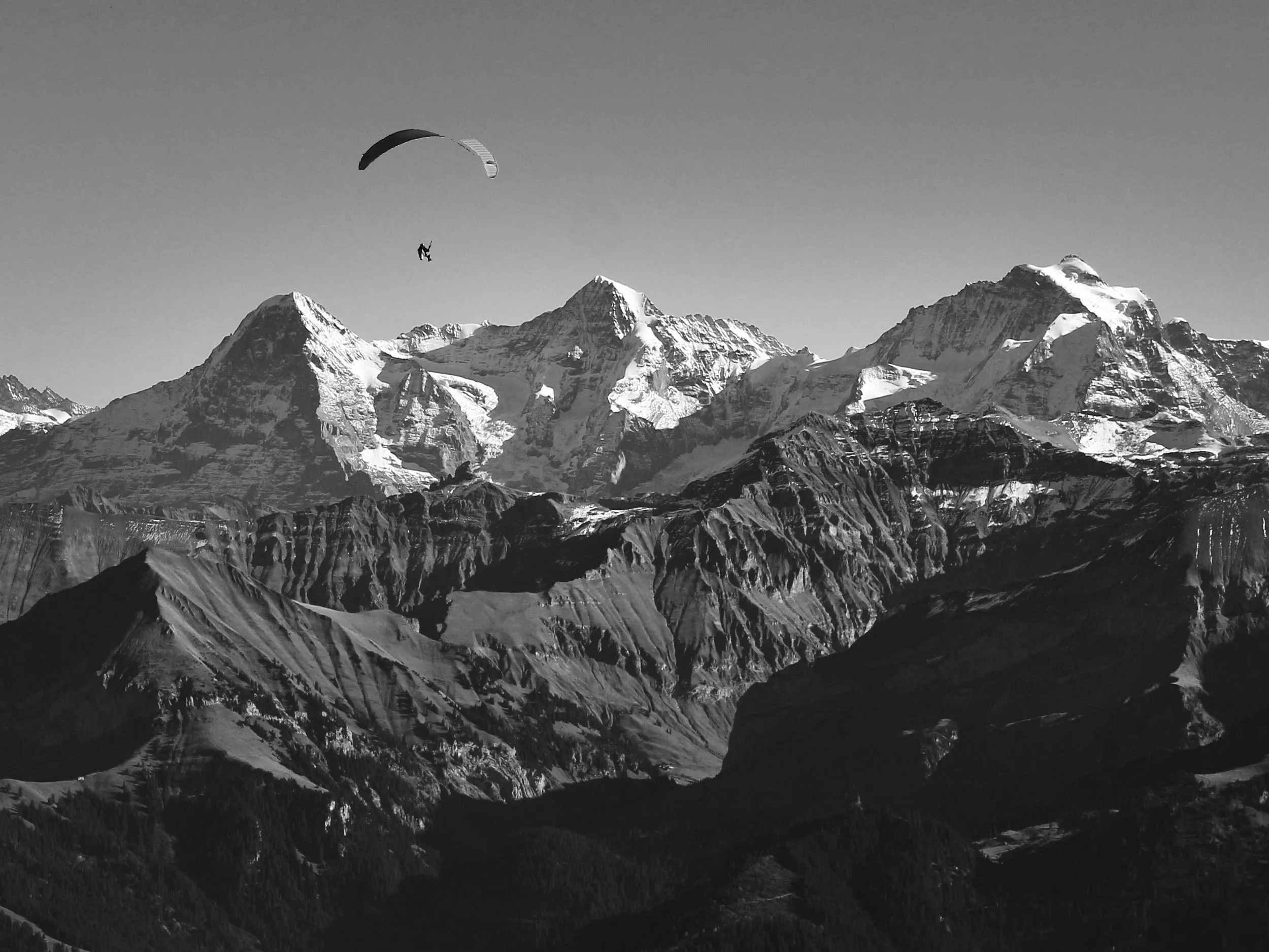 Ein Gleitschirm schwebt hoch über dramatischen, schneebedeckten Berggipfeln und schroffen Klippen unter einem klaren Himmel, der tiefe Schatten über die Landschaft wirft.