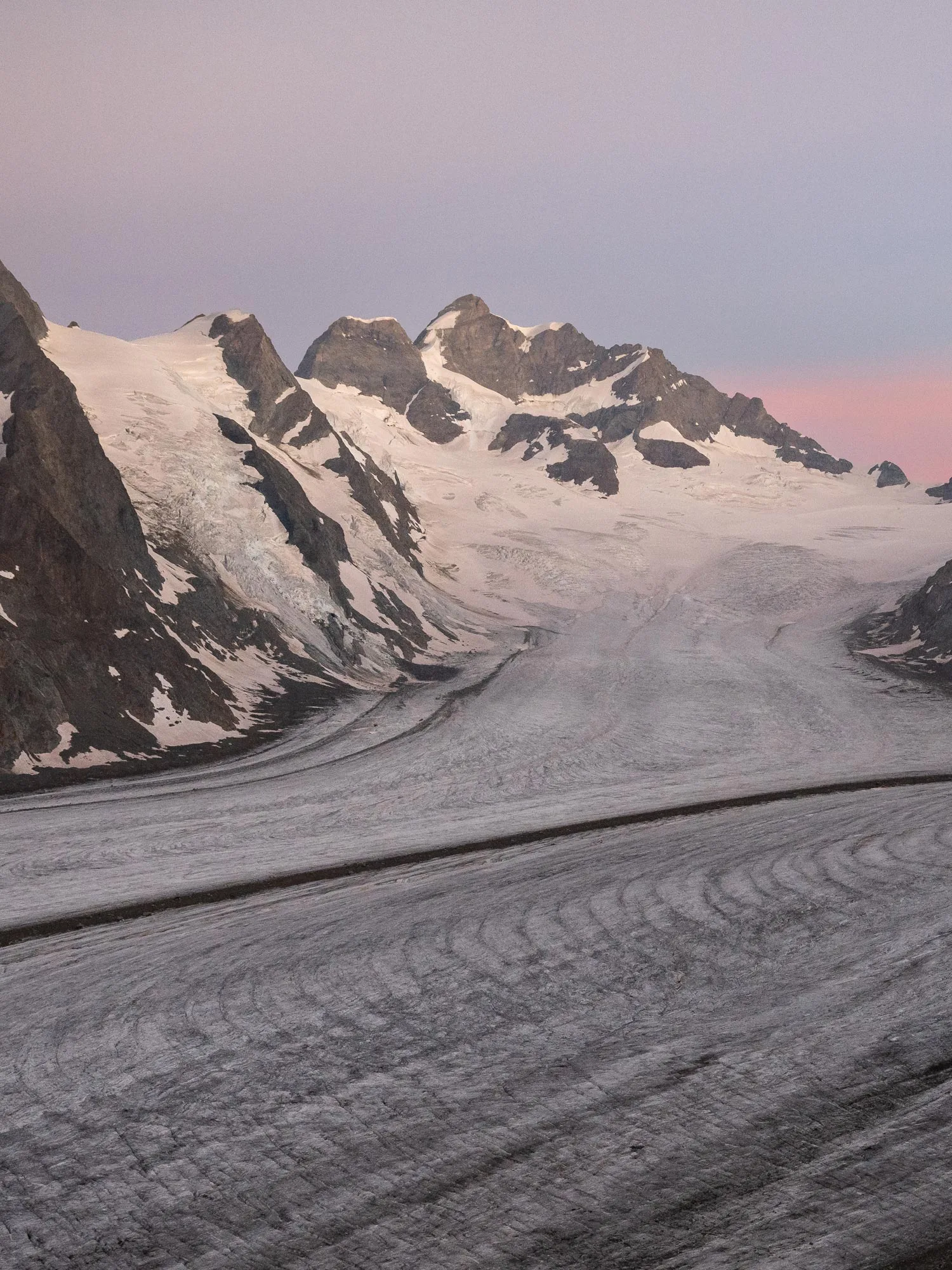 In der Abenddämmerung erheben sich schneebedeckte Berge über einem breiten, geschwungenen Gletscher. Der Himmel ist in rosa und violetten Farbtönen gehalten und dunkle Felshänge rahmen die eisige Landschaft ein.