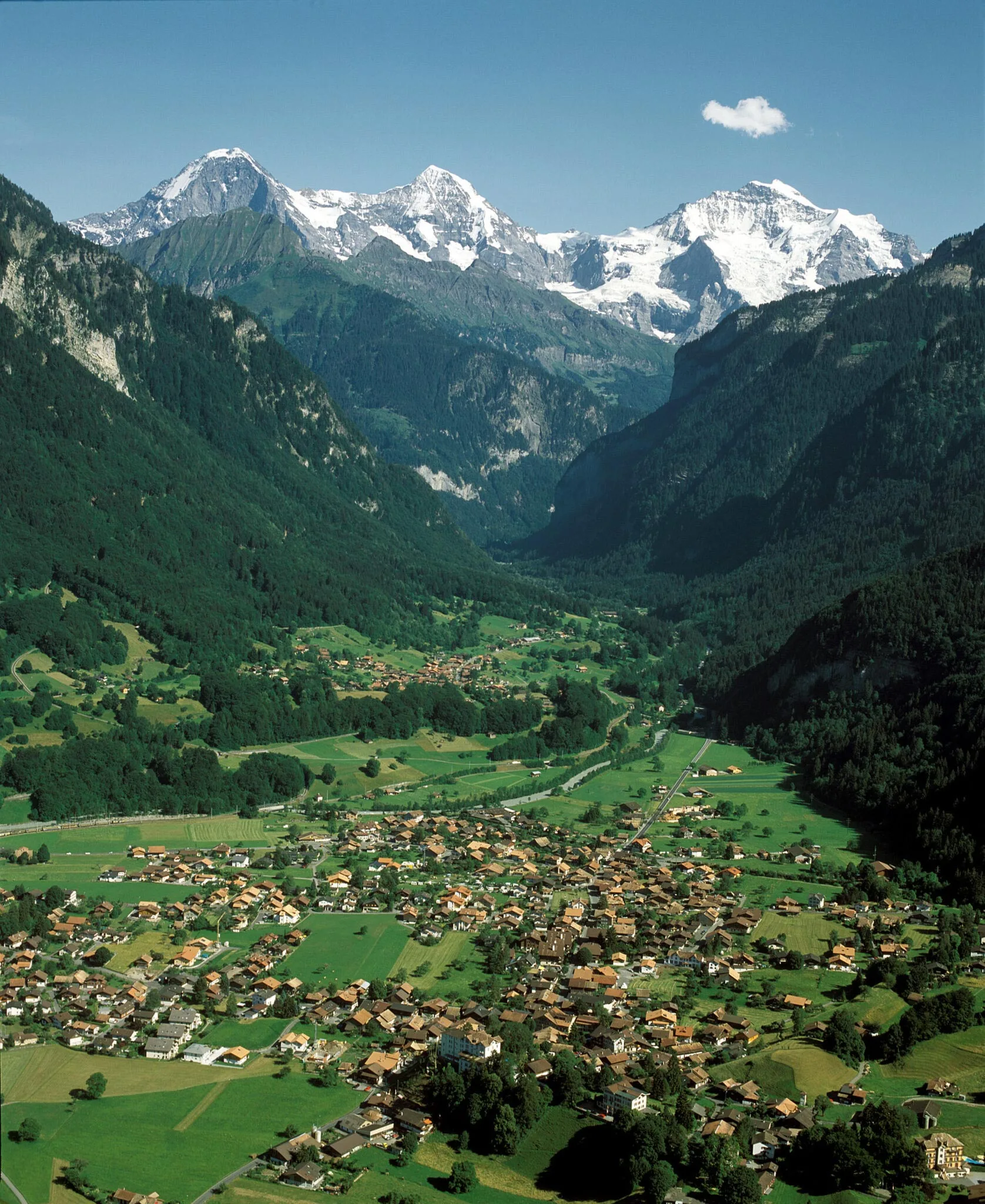 Ein malerisches Bergdorf, eingebettet in ein üppig grünes Tal, umgeben von Wäldern und dramatischen, schneebedeckten Bergen unter einem klaren blauen Himmel.