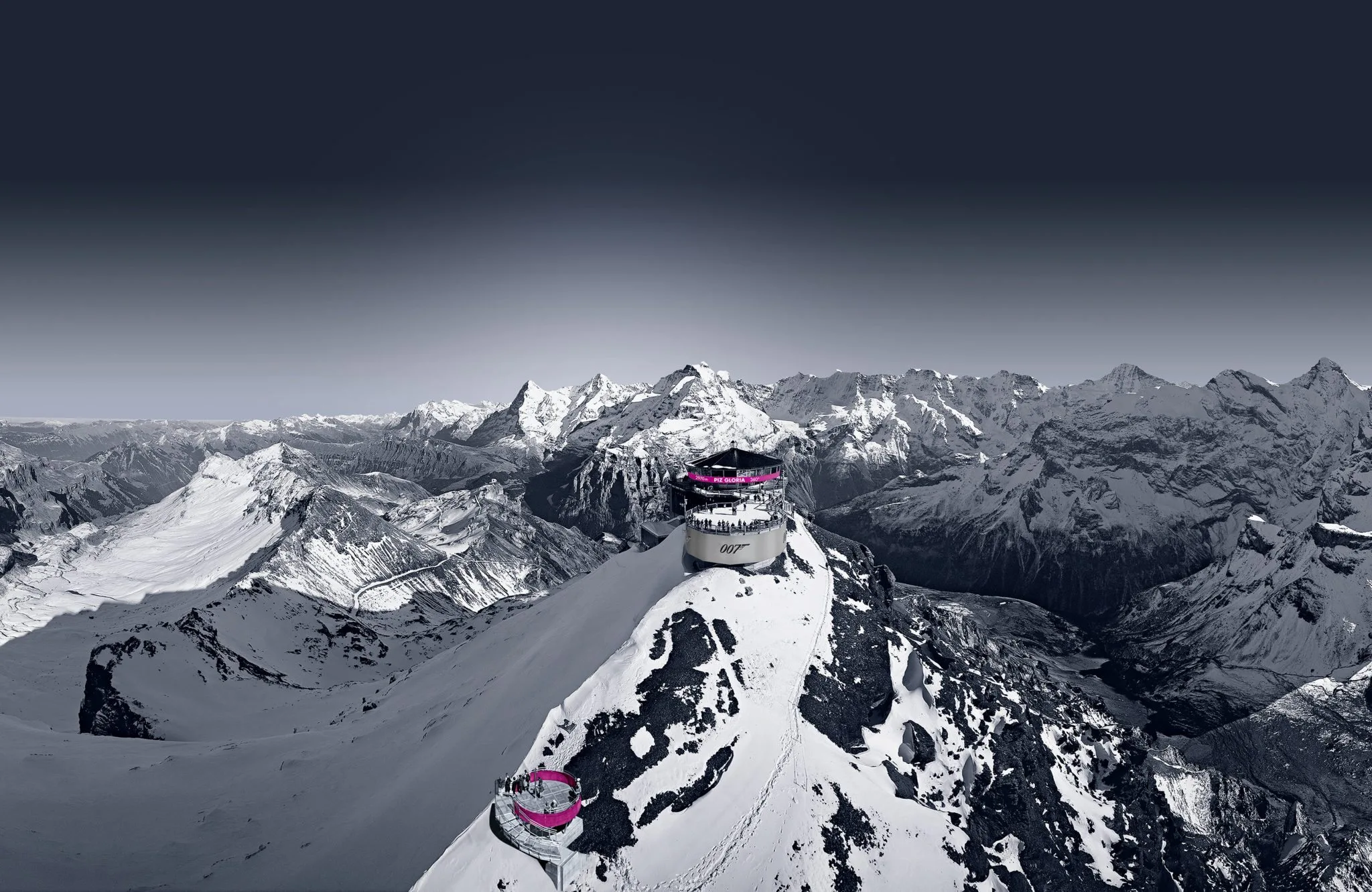 Ein futuristisches, rundes Gebäude mit rosa Akzenten steht auf einem schneebedeckten Berggipfel, umgeben von dramatischen, schneebedeckten Bergketten unter einem dunklen, klaren Himmel.