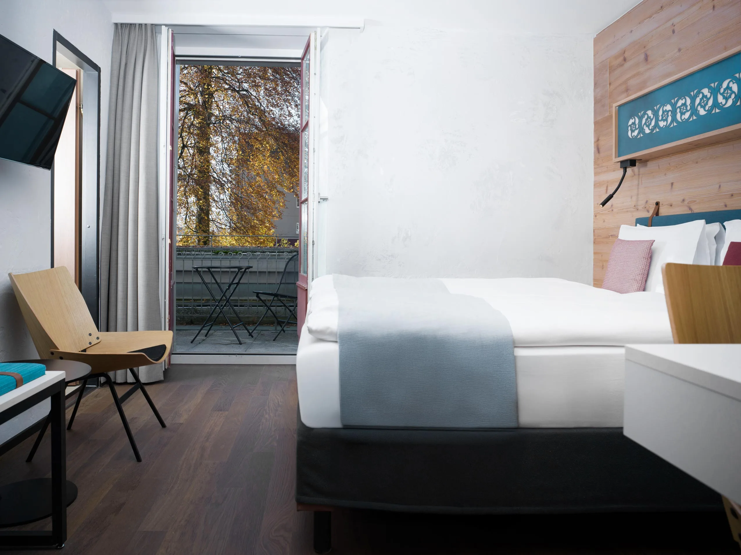 Ein modernes Hotelzimmer mit einem ordentlich gemachten Bett, Holzstühlen, einem an der Wand montierten Fernseher und einer offenen Tür, die auf einen Balkon mit einem kleinen Tisch, Stühlen und Bäumen mit sichtbaren Herbstblättern führt.