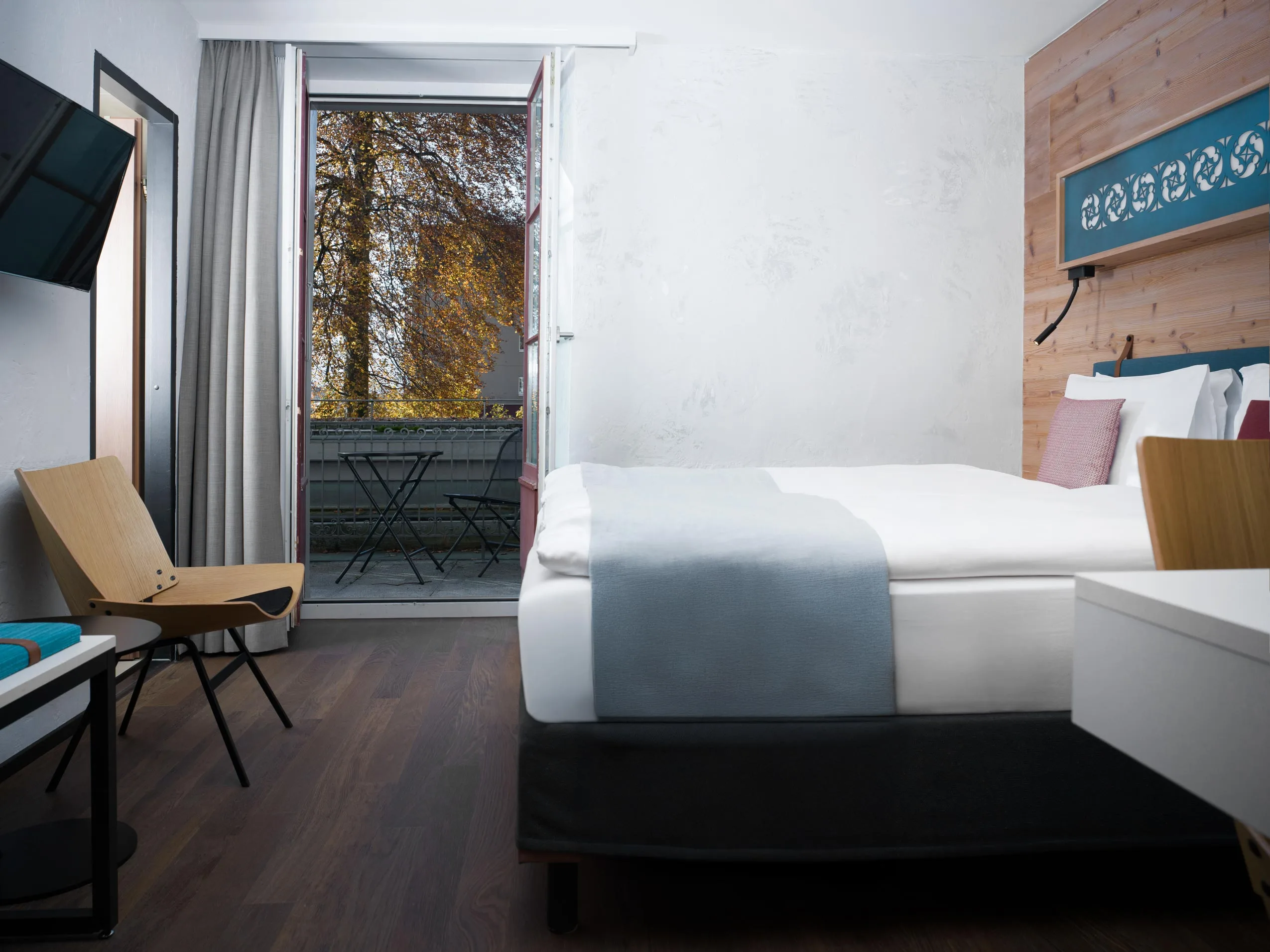 Ein modernes Hotelzimmer mit einem ordentlich gemachten Bett, Holzstühlen und einem an der Wand montierten Fernseher. Glastüren öffnen sich zu einem kleinen Balkon mit Tisch und Stühlen und bieten Aussicht auf Bäume mit Herbstlaub.