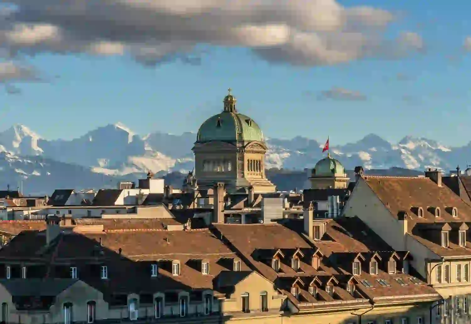 Dächer von Bern, Schweiz, mit der Kuppel des Bundeshauses in der Mitte, vor der Kulisse der schneebedeckten Alpen unter einem blauen Himmel mit vereinzelten Wolken.