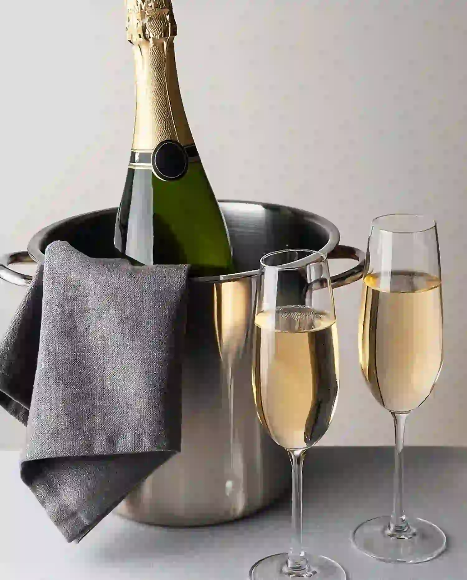 Eine Flasche Champagner in einem Metall-Eiskübel mit einem grauen Tuch darüber, daneben zwei gefüllte Champagnergläser auf einer hellgrauen Fläche – perfekt für unvergessliche Verwöhnpakete-Momente.