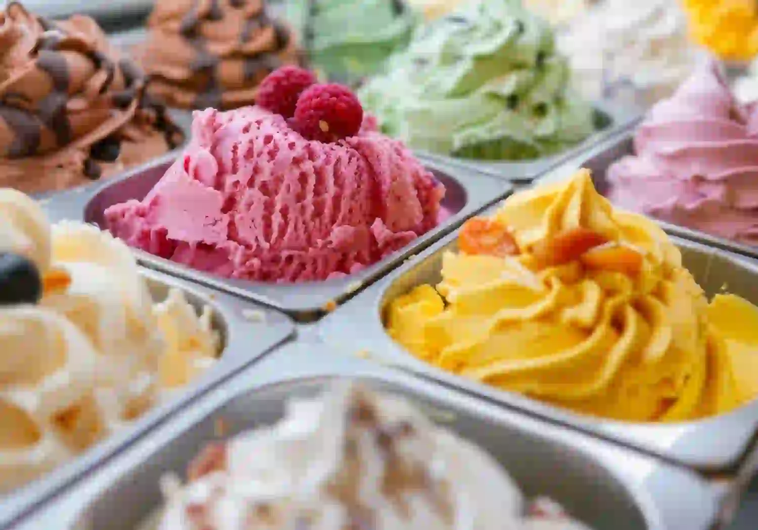 Verschiedene bunte Eissorten in Metallschalen, mit einer Nahaufnahme von rosa Himbeer-Eis mit frischen Himbeeren obendrauf und einem gelben Eis mit Nussstückchen im Vordergrund.