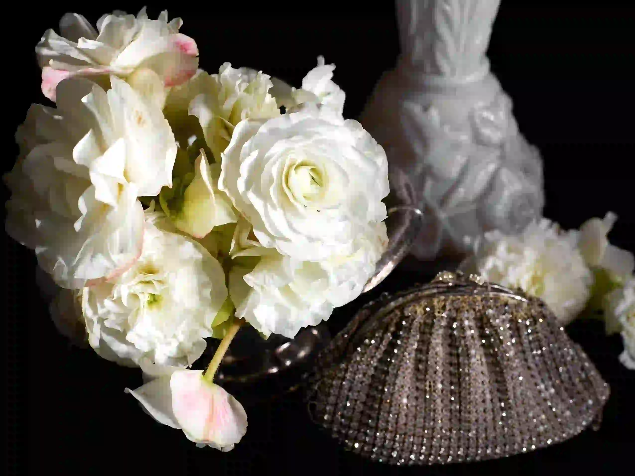 Eine Nahaufnahme weißer Rosen in einer silbernen Vase neben einer Clutch mit Perlen und einer weißen Dekovase vor einem schwarzen Hintergrund – ein elegantes Arrangement, perfekt für Verwöhnpakete und luxuriöse Umgebungen.