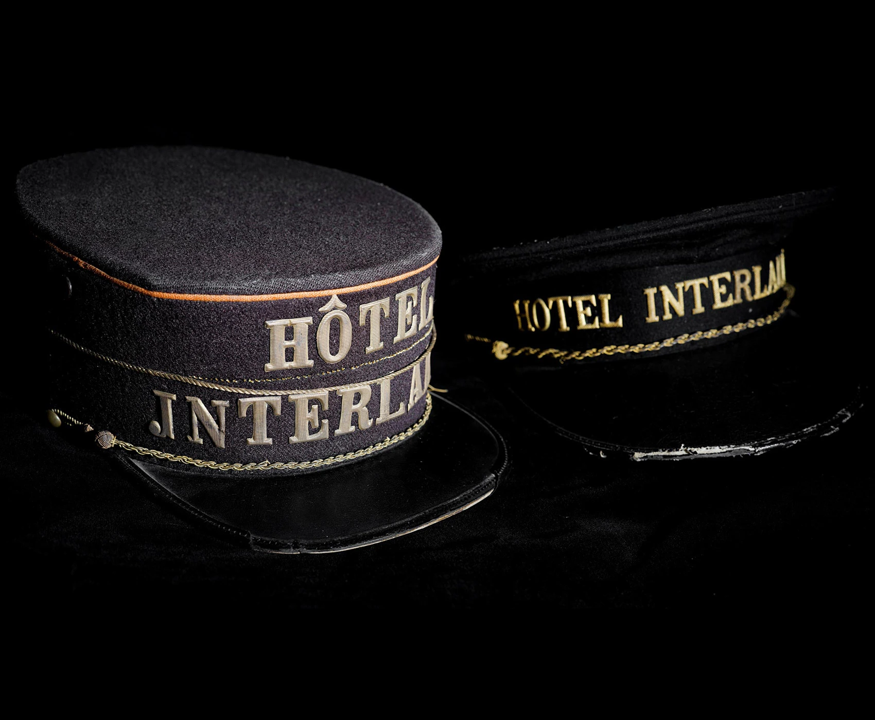Zwei schwarze Hotelportiermützen im Vintage-Stil mit goldgeflochtenem Besatz und gesticktem Text mit der Aufschrift „Hôtel Interlaken“ und „Hotel Interlaken“, abgebildet auf schwarzem Hintergrund.