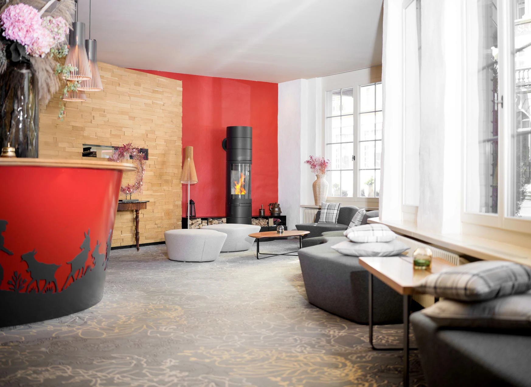 Eine moderne Hotellobby mit roten Akzenten, einem geschwungenen Rezeptionstresen mit Tiersilhouetten, einer holzgetäfelten Wand, einem schwarzen Kamin, gepolsterten Sitzgelegenheiten und großen Fenstern, die natürliches Licht hereinlassen.