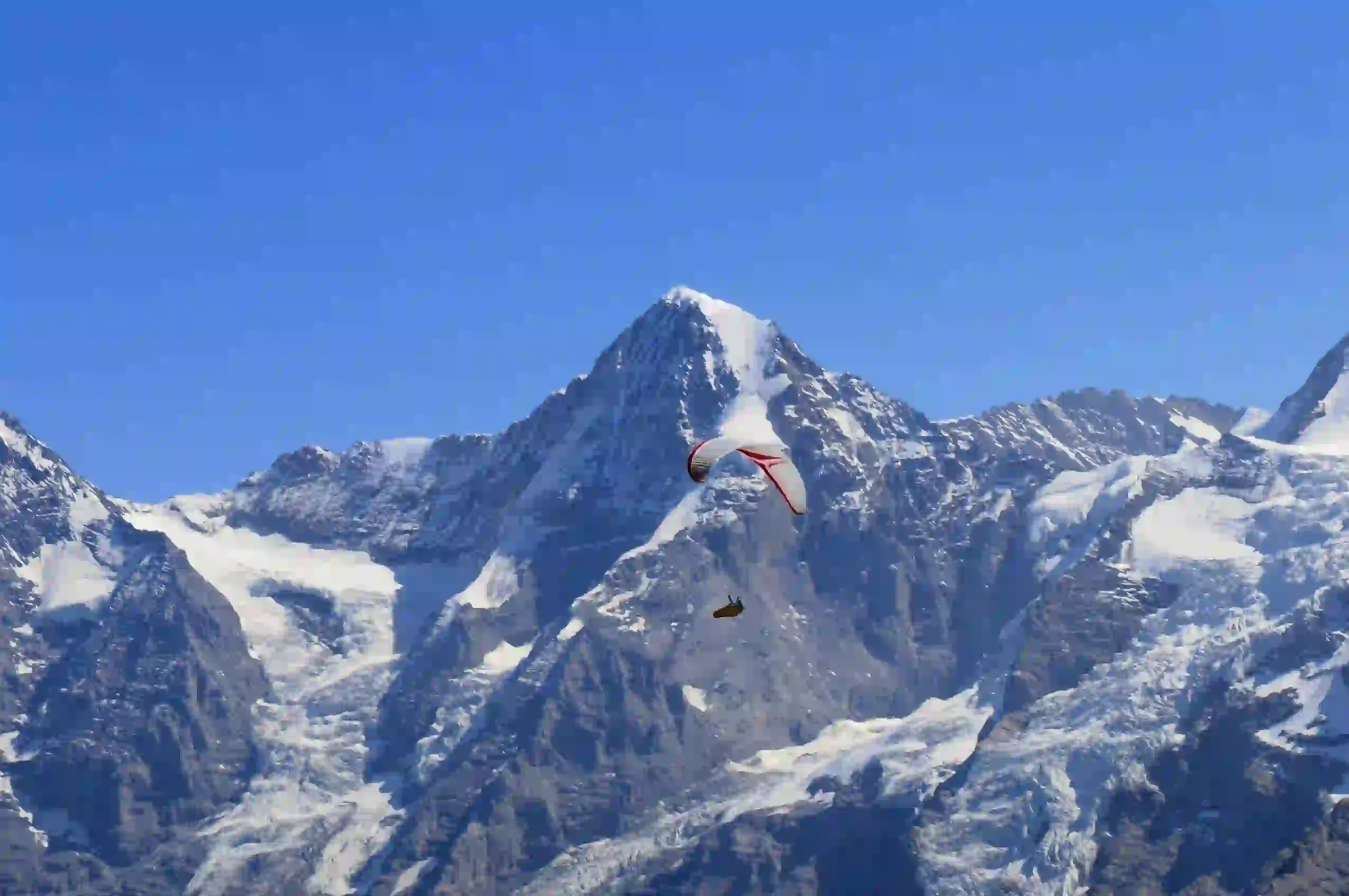 Ein Gleitschirm schwebt vor hohen, schneebedeckten Bergen vor einem klaren blauen Himmel, im Hintergrund sind schroffe Gipfel und Gletscher zu sehen.